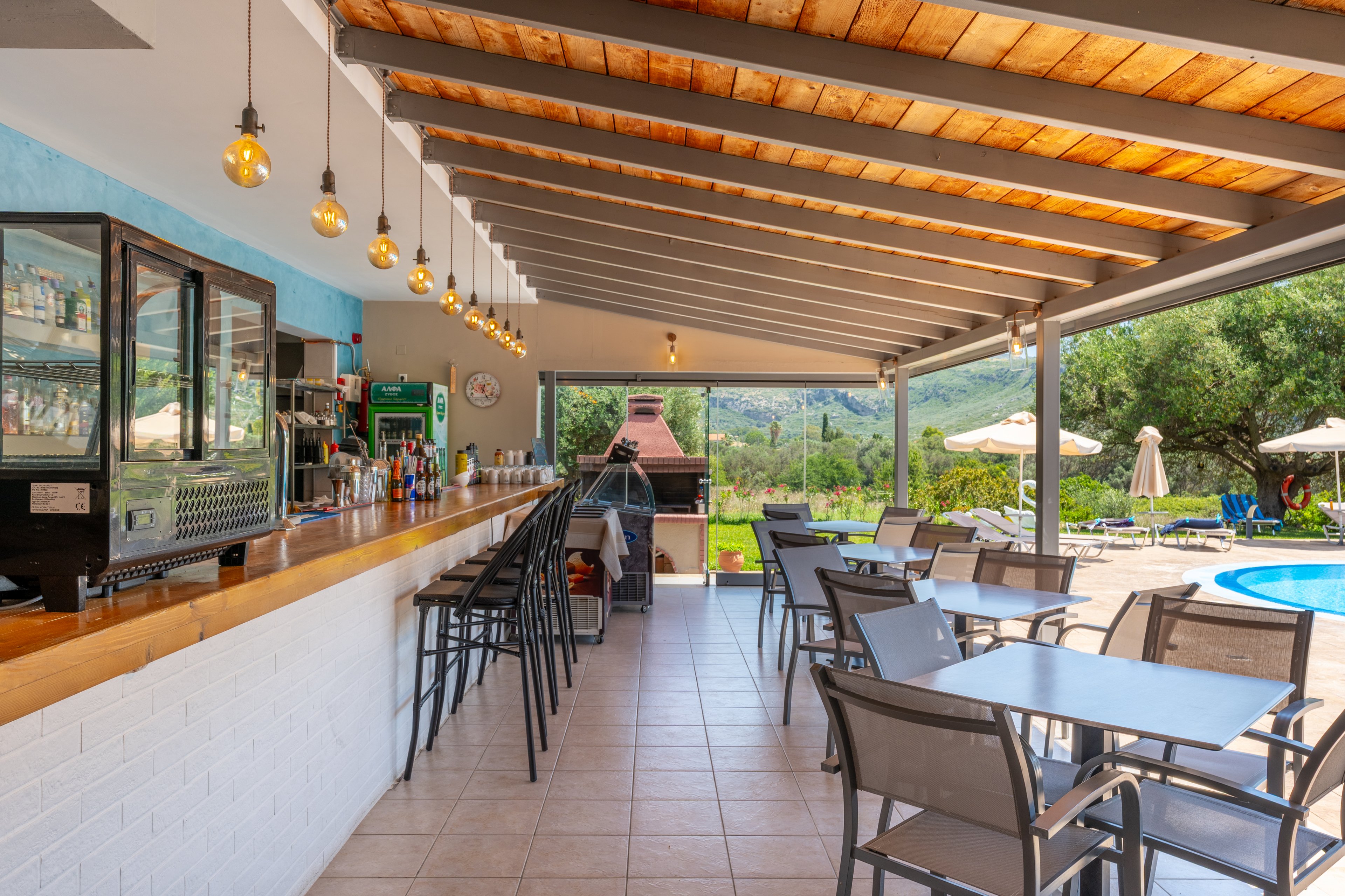 An open-air bar with a long wooden counter and high stools overlooks a poolside terrace. Modern metal tables and chairs are arranged on tiled flooring under a slatted wooden roof with hanging pendant lights. A glass-fronted fridge and various bottles are visible behind the bar, while a brick grill stands at the far end. In the background, sun loungers and umbrellas sit beside a bright blue swimming pool surrounded by greenery and hills.