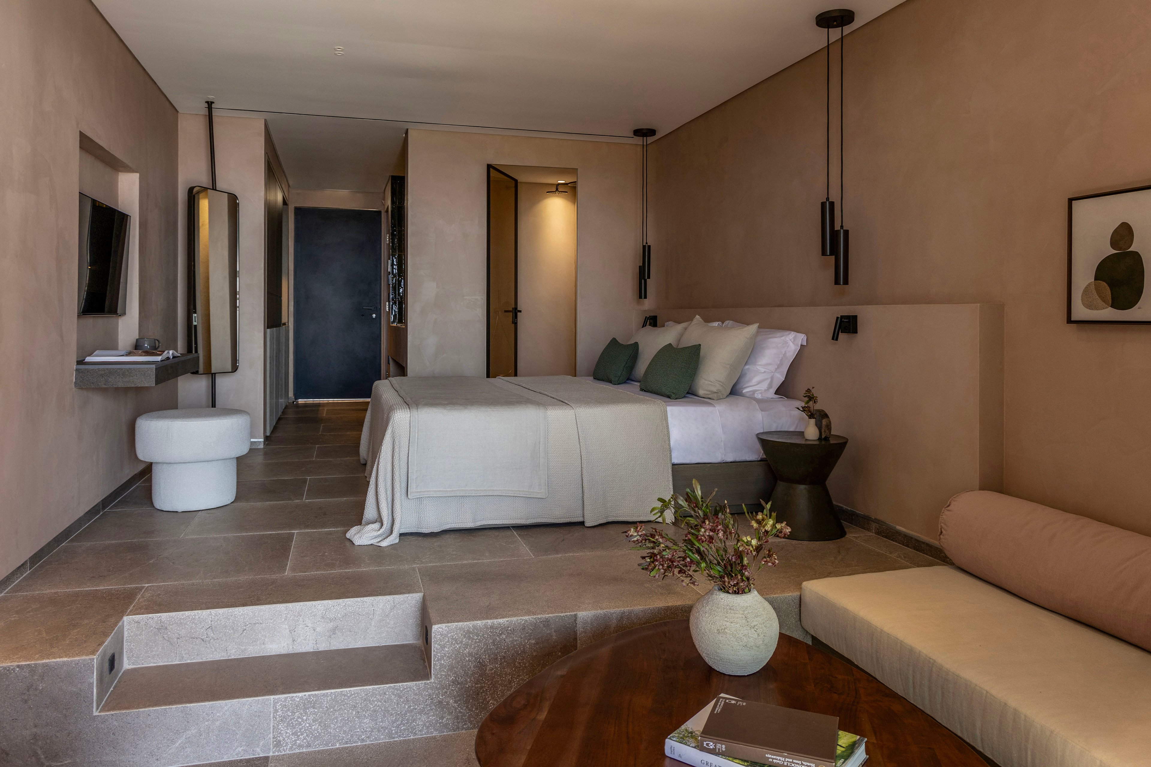 Contemporary hotel room featuring a neatly made double bed with neutral bedding and accent pillows. The space includes a small seating area with a cushioned bench and round wooden table holding a vase with greenery and books. Warm beige walls, stone flooring, and recessed lighting create a cozy, minimalist atmosphere. A wall-mounted television, modern side tables, and simple artwork complete the stylish interior.