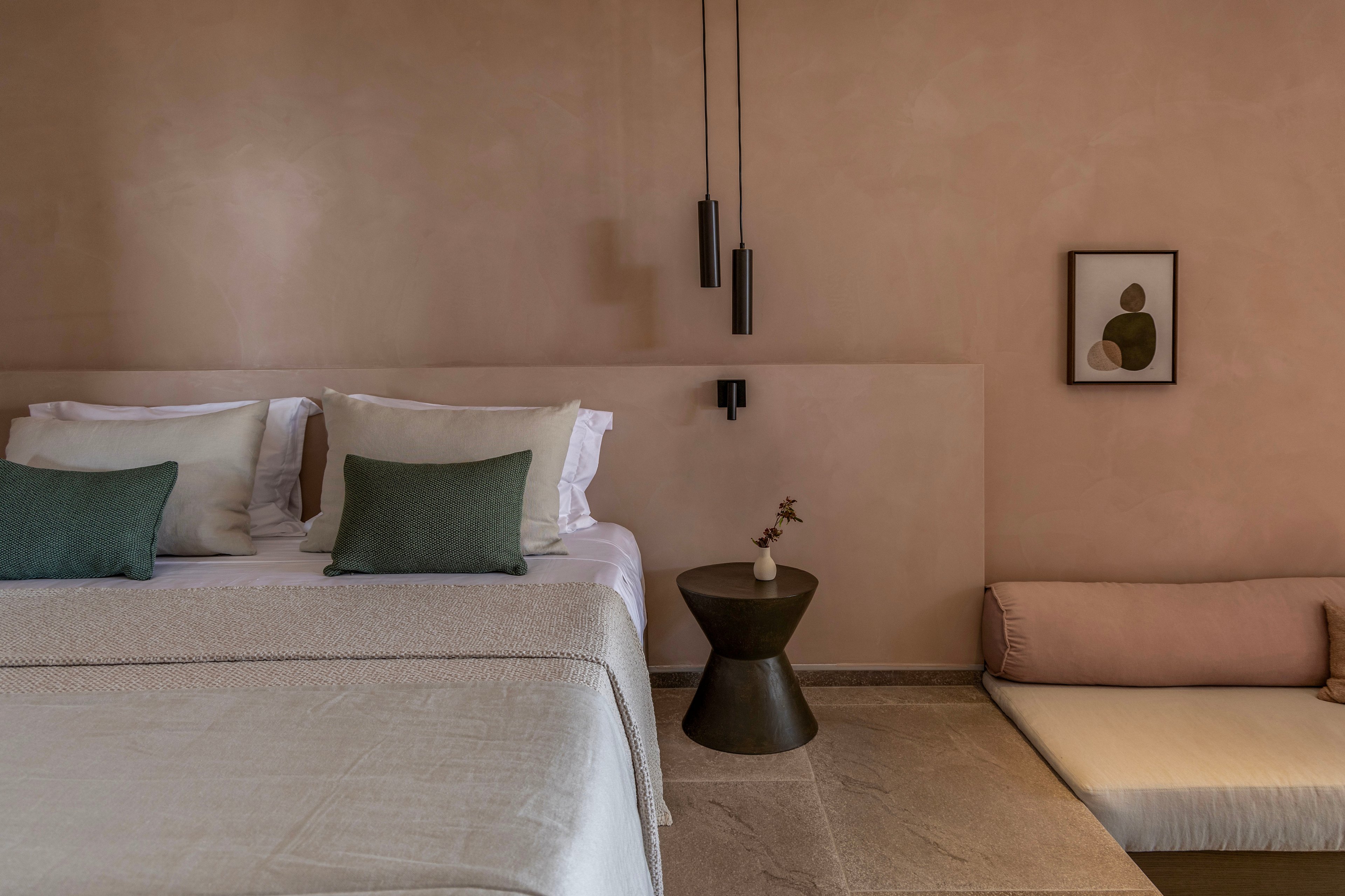 A minimalist bedroom features a neatly made bed with white linens and green accent pillows against a smooth beige wall. Beside the bed stands a small dark side table holding a simple vase with dried flowers. Modern black pendant lights hang above, adding vertical interest to the calm space. A low cushioned bench and a framed abstract artwork complete the serene, contemporary interior.