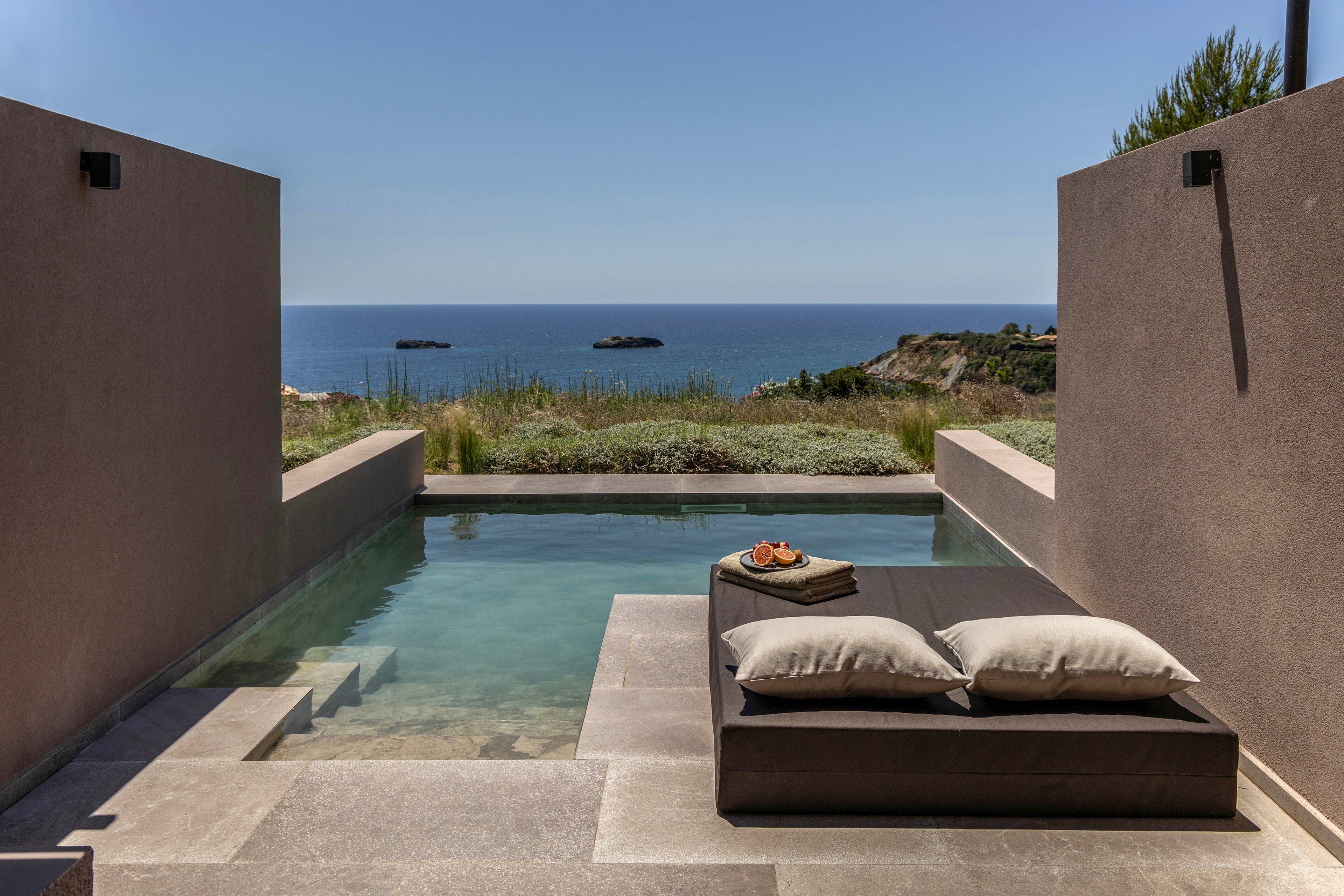 A sleek private plunge pool stretches toward a distant, calm sea under a clear blue sky. Minimalist stone walls frame the view, leading the eye to the horizon and small rocky islets offshore. In the foreground, a low sunbed with neutral cushions and a tray of fruit suggests relaxation and resort luxury. Natural grasses soften the transition between the contemporary terrace and the coastal landscape, creating a serene, upscale retreat atmosphere.