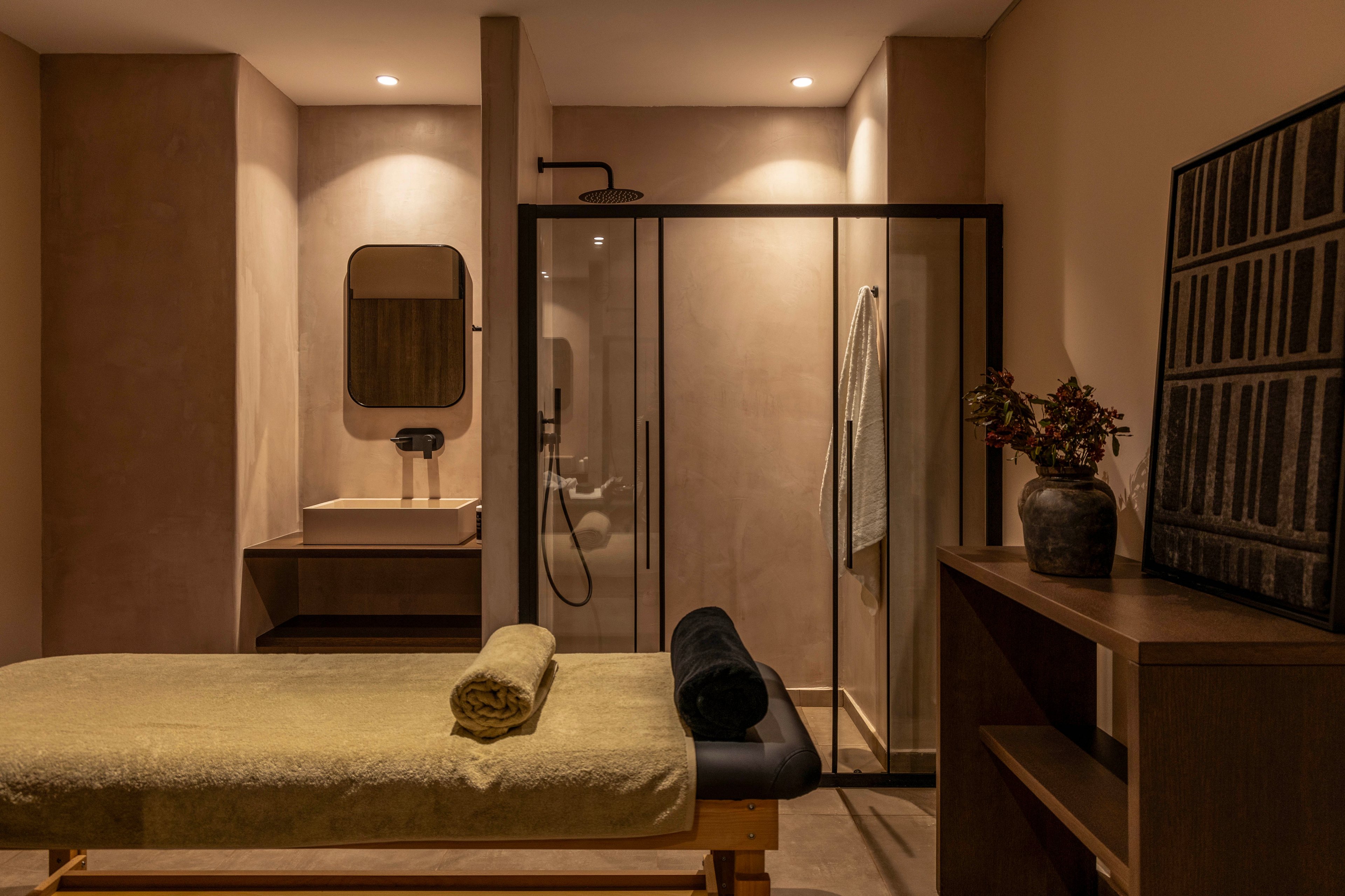 A contemporary spa treatment room features a massage table with neatly rolled towels in warm, neutral tones. In the background, a glass-enclosed walk-in shower and a minimalist vanity with mirror create a clean, modern look. Soft, recessed lighting and beige walls contribute to a calm, intimate atmosphere. A wooden shelf with a decorative vase and framed artwork adds subtle, stylish accents to the space.