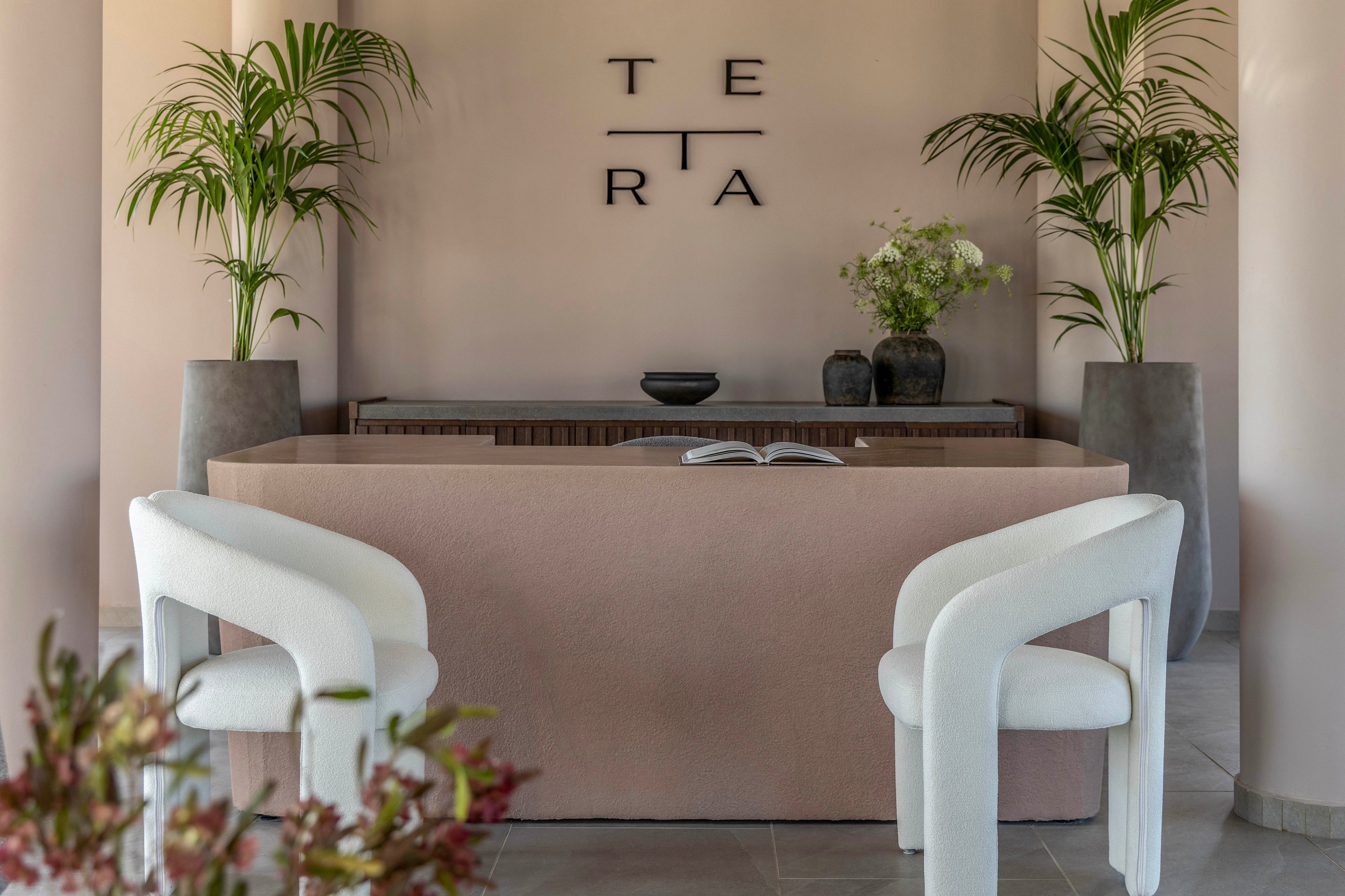 A contemporary reception area features a soft pink counter with an open book placed on top. Two sculptural white armchairs face the desk, creating a welcoming seating arrangement. Tall potted plants frame the space, adding greenery against the neutral walls. On the back wall, the word "TERA" is displayed in bold black letters, enhancing the modern, minimalist aesthetic.
