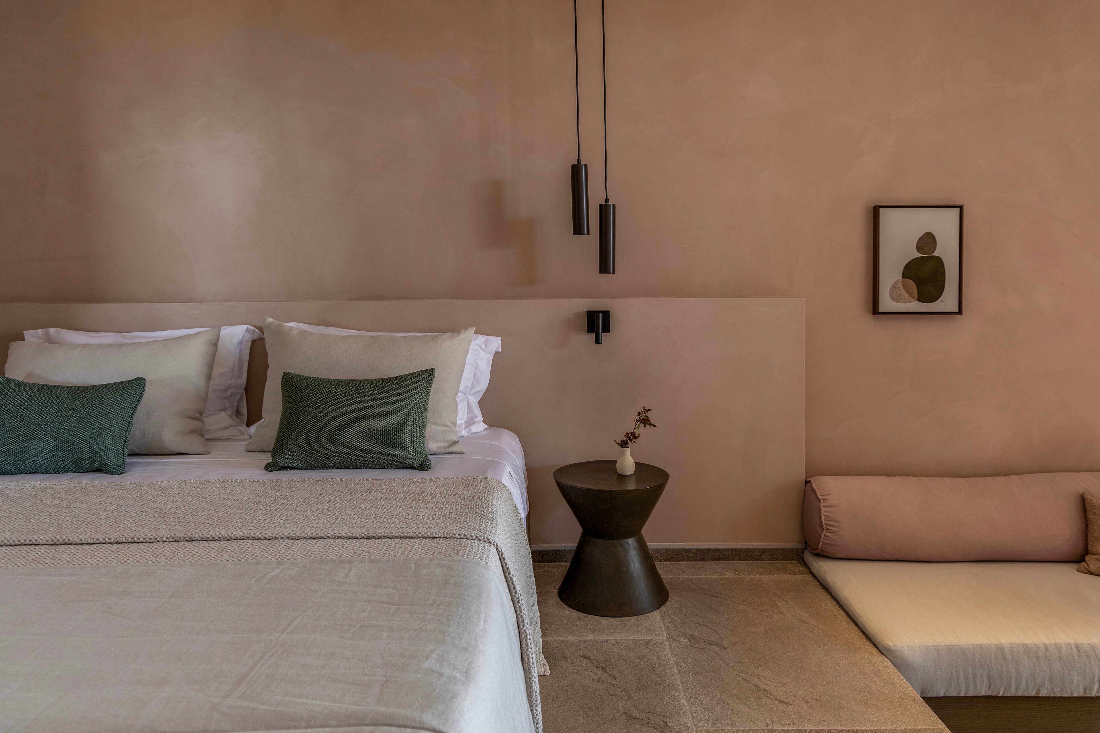 A contemporary bedroom features a neatly made bed with neutral bedding and two green accent pillows. The space is designed with warm, earthy-toned walls and a low built-in seating area with a blush cushion. A small sculptural side table holds a simple vase with dried flowers, while slim pendant lights hang above. A framed abstract artwork adds a subtle artistic touch to the minimalist interior.