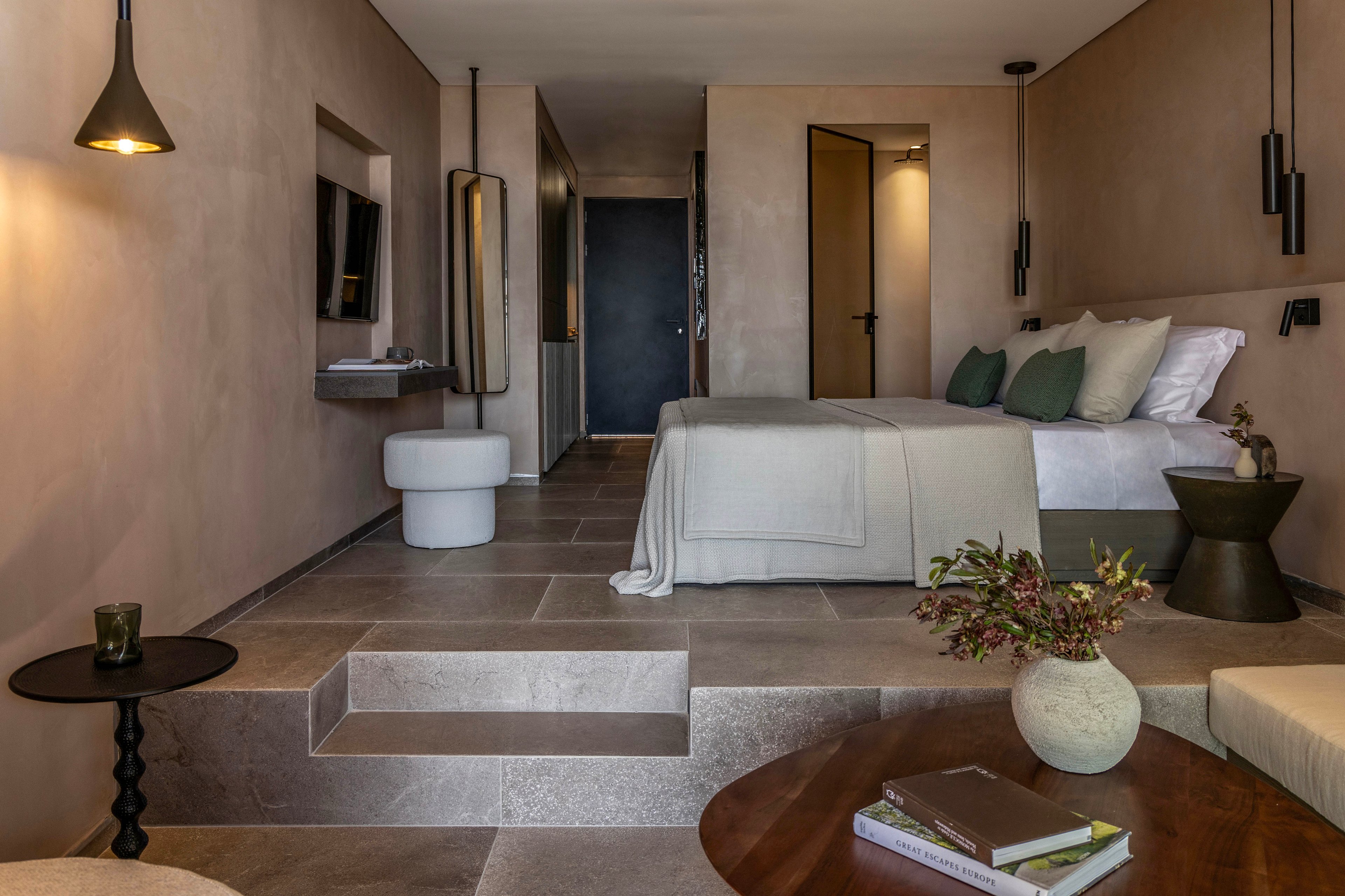A contemporary hotel room features a neatly made bed with white linens and green accent pillows. The space is designed with large stone floor tiles, soft beige walls, and built-in steps leading to the sleeping area. Modern pendant lighting, a wall-mounted television, and minimalist furniture including stools and side tables enhance the clean aesthetic. A round wooden table in the foreground holds books and a decorative vase with foliage, adding warmth and texture to the serene interior.