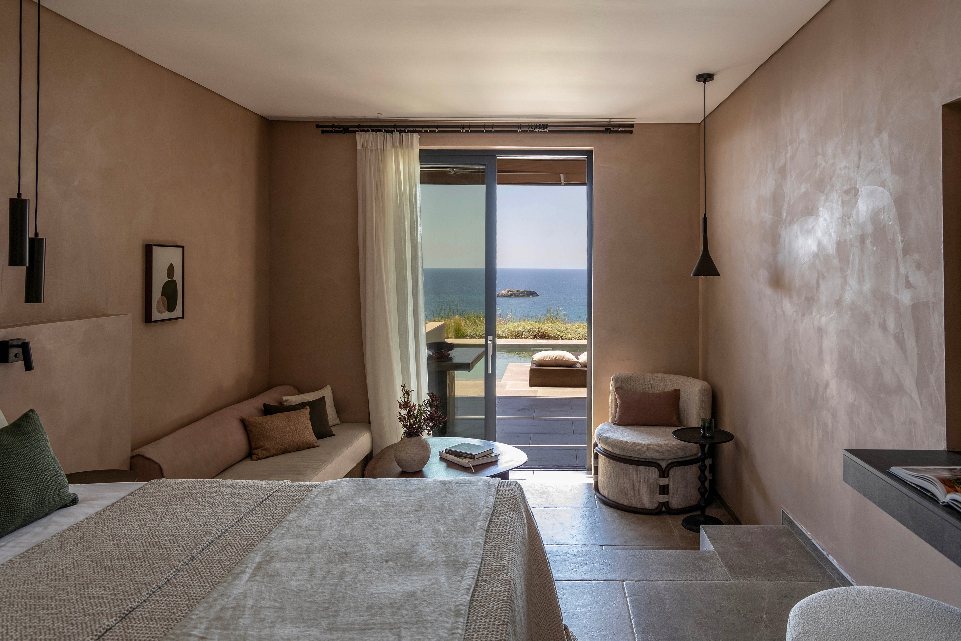 A contemporary hotel room features a neutral-toned bed and a small seating area with armchair and side table. Large sliding glass doors open to a terrace overlooking the ocean and a distant rocky islet. Soft natural light filters through sheer curtains, highlighting the warm beige walls and modern pendant lighting. The overall atmosphere is calm, minimalist, and luxurious, ideal for a relaxing seaside stay.
