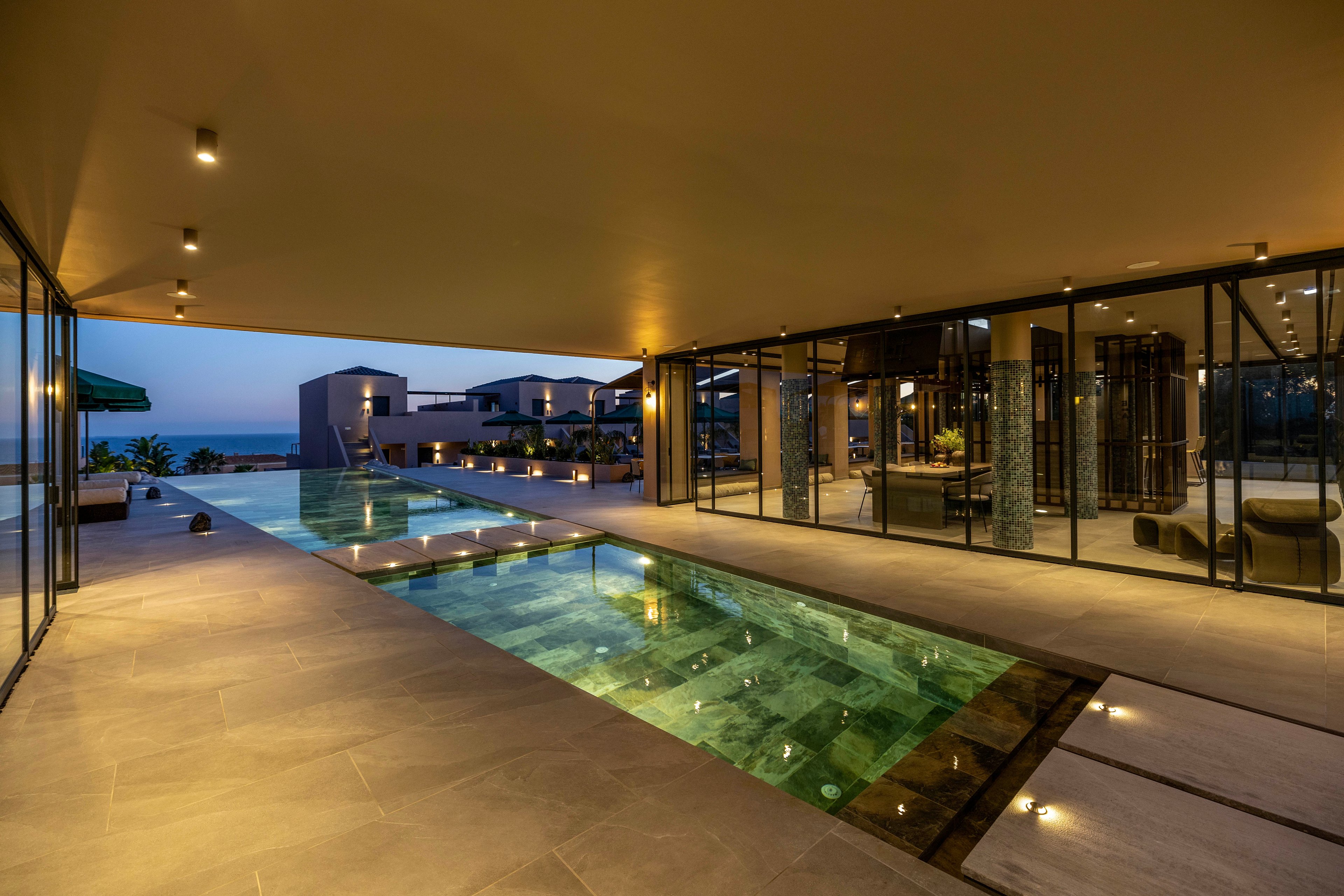 A contemporary luxury villa features a long, narrow swimming pool illuminated by warm lights at dusk. The scene is framed by sleek concrete flooring, glass walls, and a covered terrace with recessed ceiling lights. In the background, modern residential structures overlook a calm coastal horizon under a deep blue evening sky. The overall atmosphere is tranquil and sophisticated, emphasizing modern architecture and resort-style living.