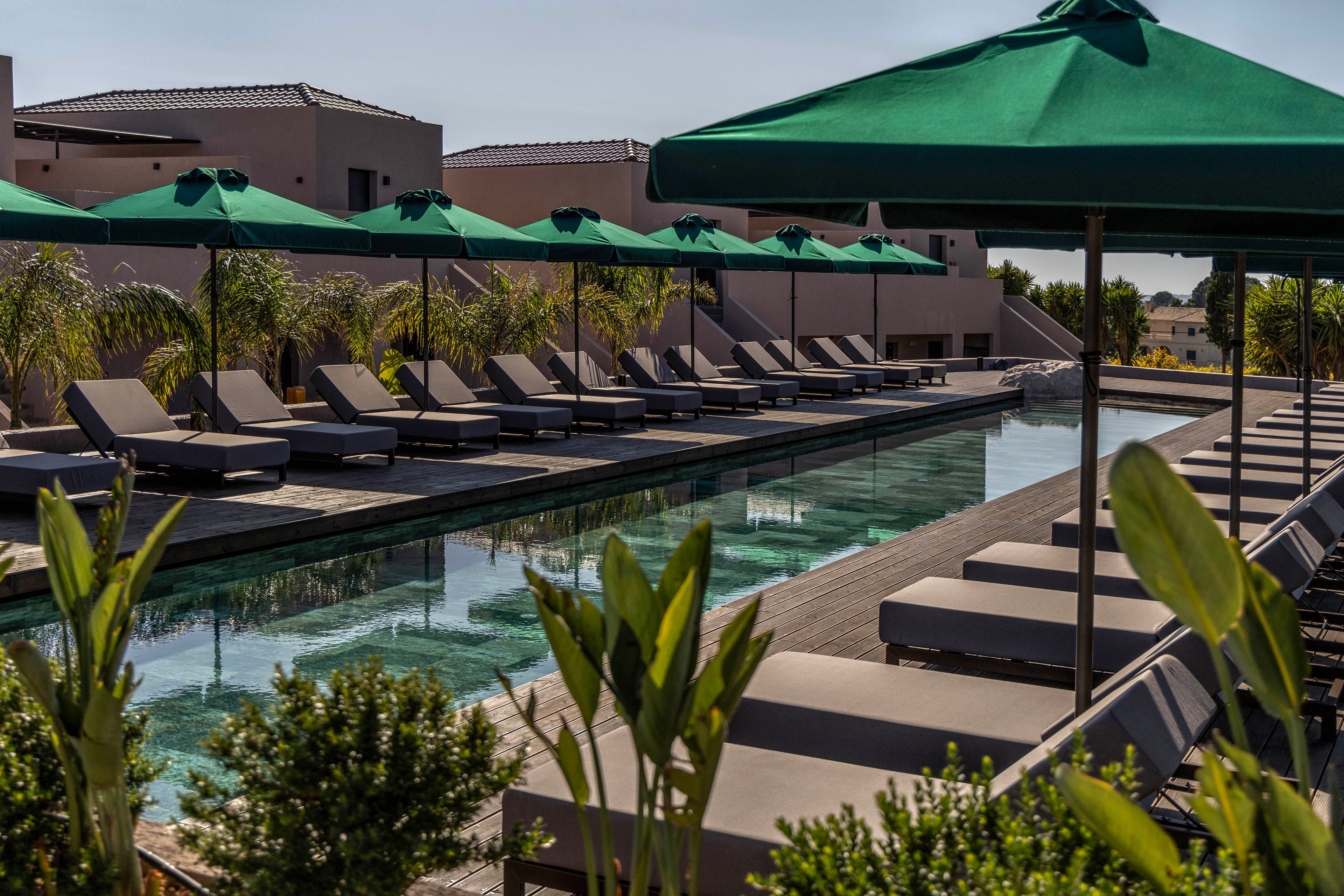 A long, narrow swimming pool is lined with cushioned sun loungers on both sides in a contemporary resort setting. Green parasols provide shade along the wooden deck, while tropical plants frame the foreground. Minimalist buildings with flat roofs and neutral tones rise behind the pool, suggesting a warm, sunny climate. The overall scene conveys relaxation, leisure, and upscale outdoor comfort.