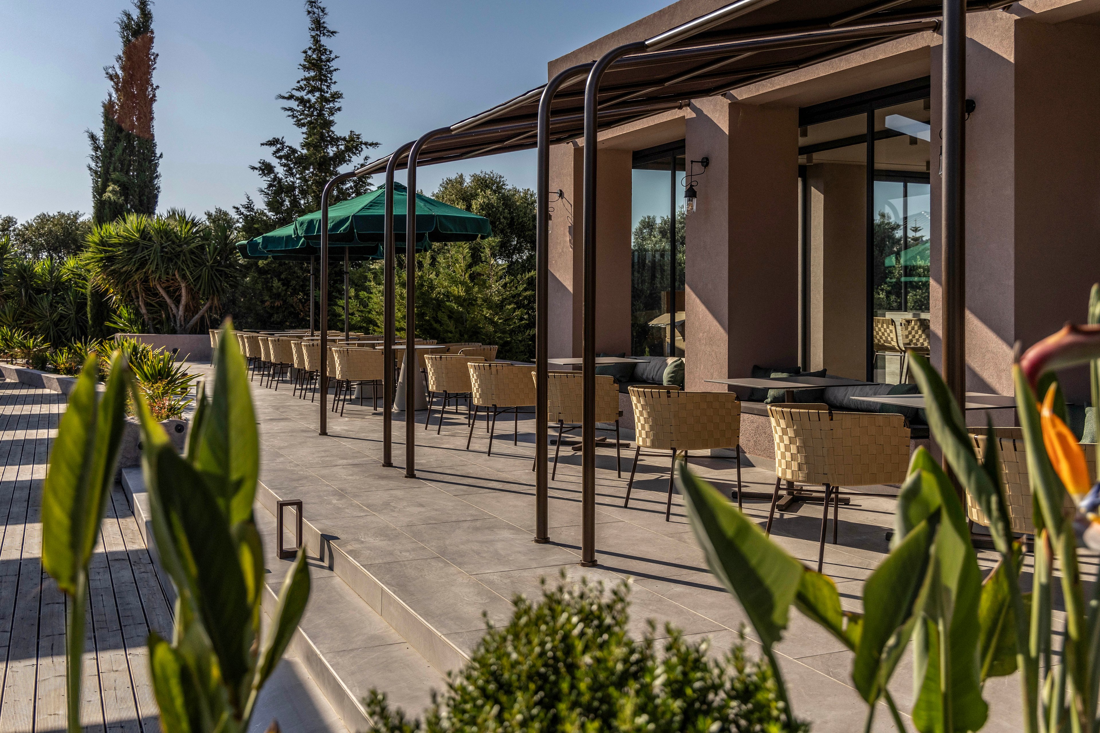 A contemporary building features a long outdoor terrace lined with cushioned chairs and tables. Metal pergola structures frame the seating area, providing shade along the walkway. Green umbrellas and lush plants surround the patio, creating a relaxed, resort-like atmosphere. Warm sunlight and clear skies highlight the modern architecture and landscaped garden.