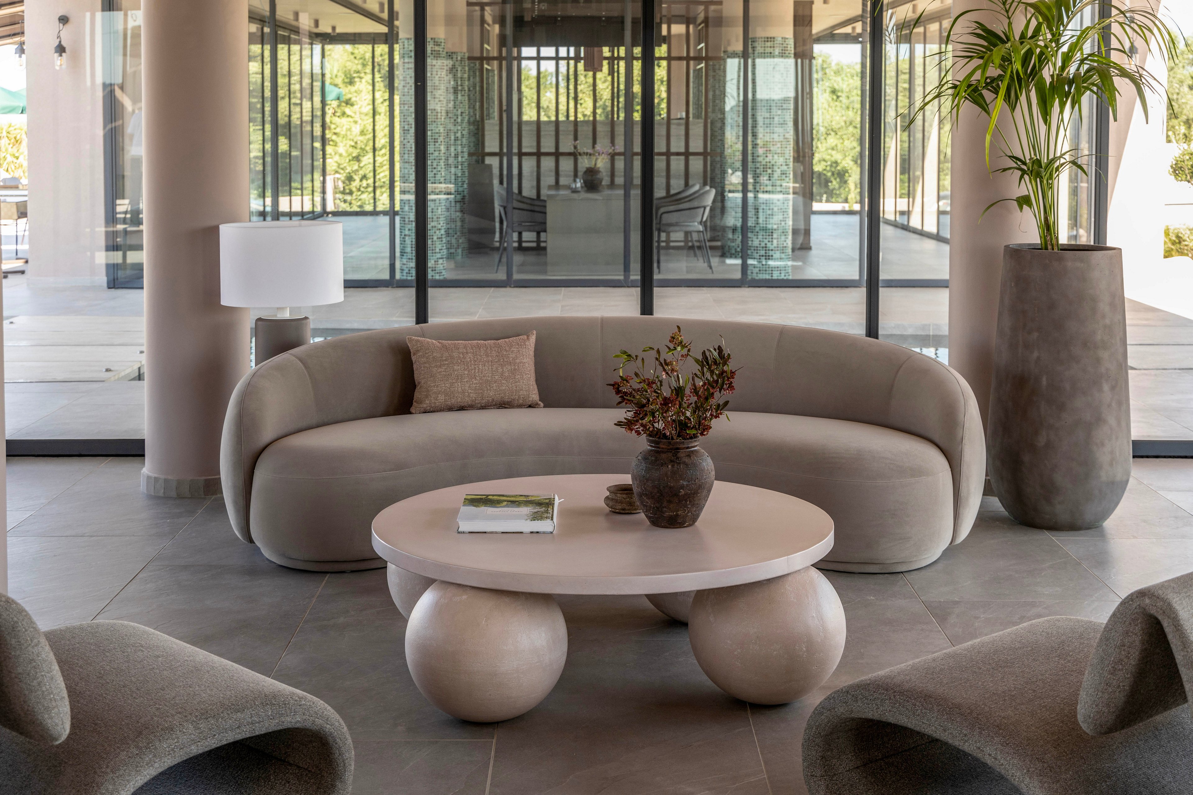 A contemporary lounge area features a curved beige sofa centered around a round coffee table with sculptural ball-shaped legs. A ceramic vase with dried foliage and a book rest on the table, adding subtle decorative accents. Matching armchairs, a floor planter with a tall green plant, and a minimalist table lamp complete the neutral-toned interior. Large glass doors in the background open to an outdoor view, enhancing the airy and sophisticated atmosphere.
