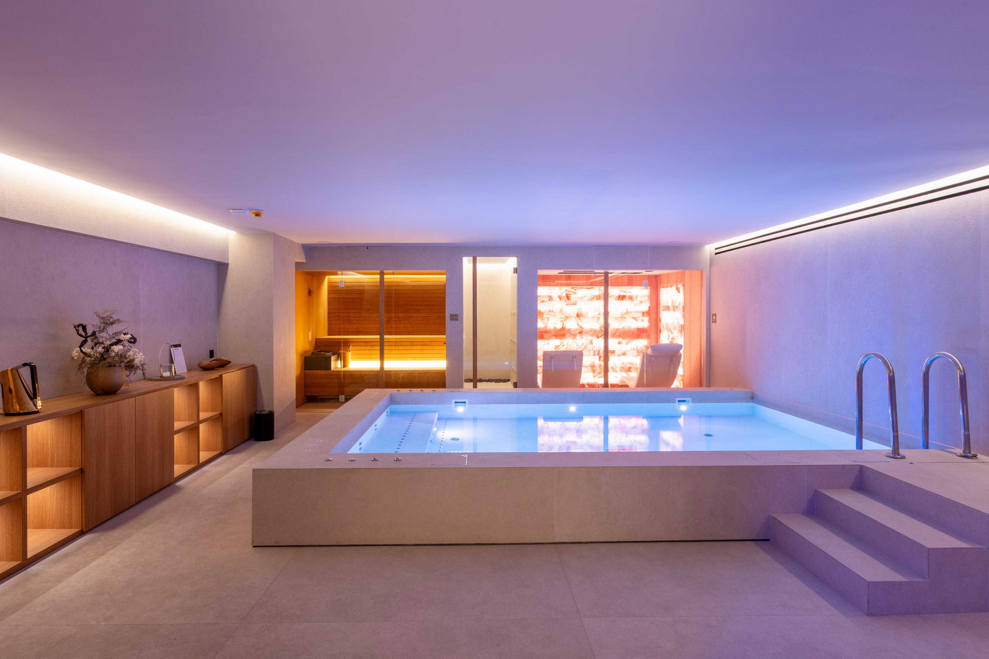 A contemporary indoor spa room features a central rectangular pool with steps and metal handrails. Warm ambient lighting illuminates a wooden sauna area and a glowing salt wall in the background. Minimalist cabinetry with decorative objects lines one side, contrasting with smooth light-colored floors and walls. The overall atmosphere is calm, luxurious, and designed for relaxation and wellness.