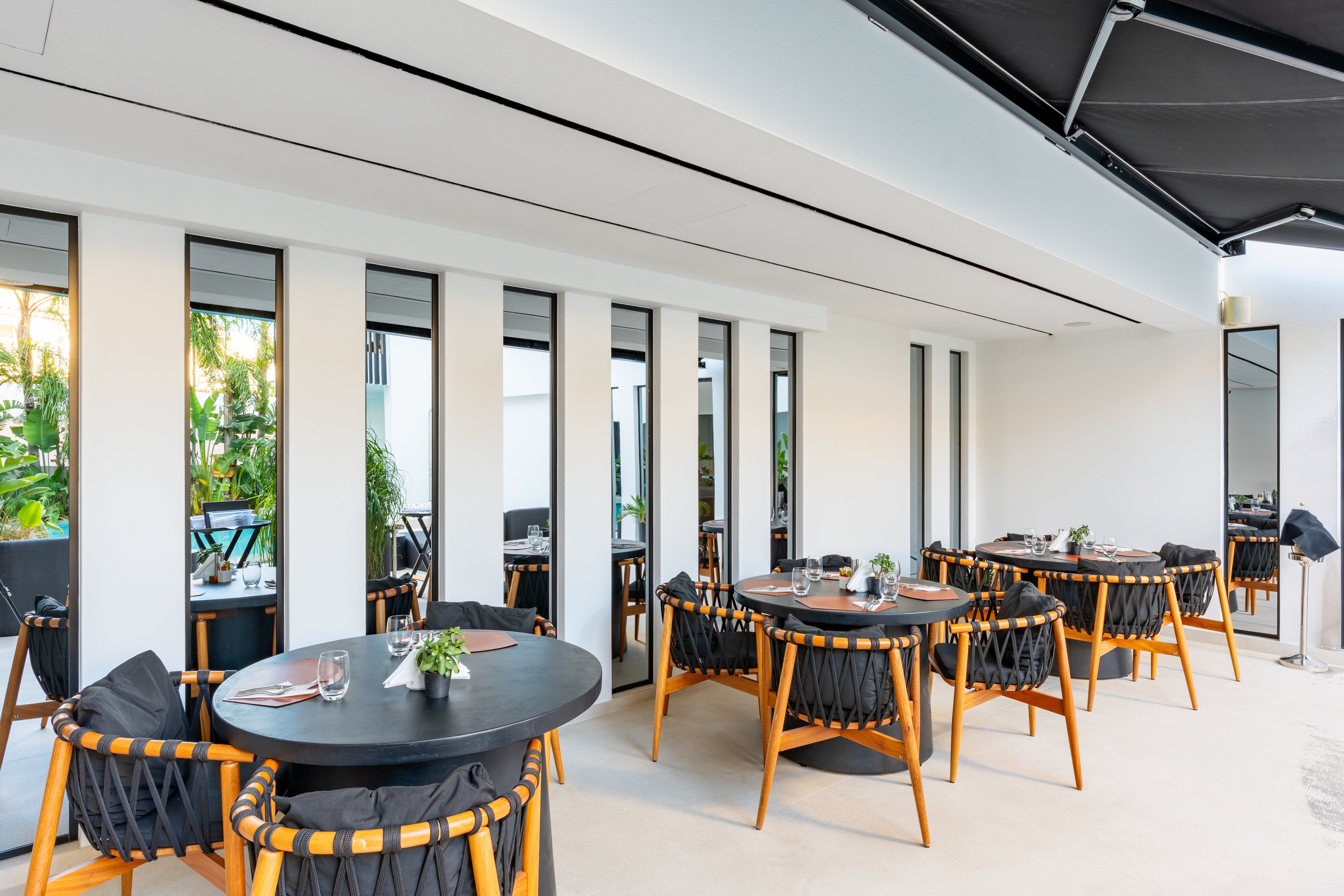 Contemporary round tables and woven chairs are arranged on a bright outdoor restaurant terrace. The setting features white walls with tall vertical windows and views of lush green plants outside. Table settings include plates, cutlery, and small floral centerpieces, suggesting readiness for guests. The overall atmosphere is clean, stylish, and relaxed, with natural light enhancing the modern design.