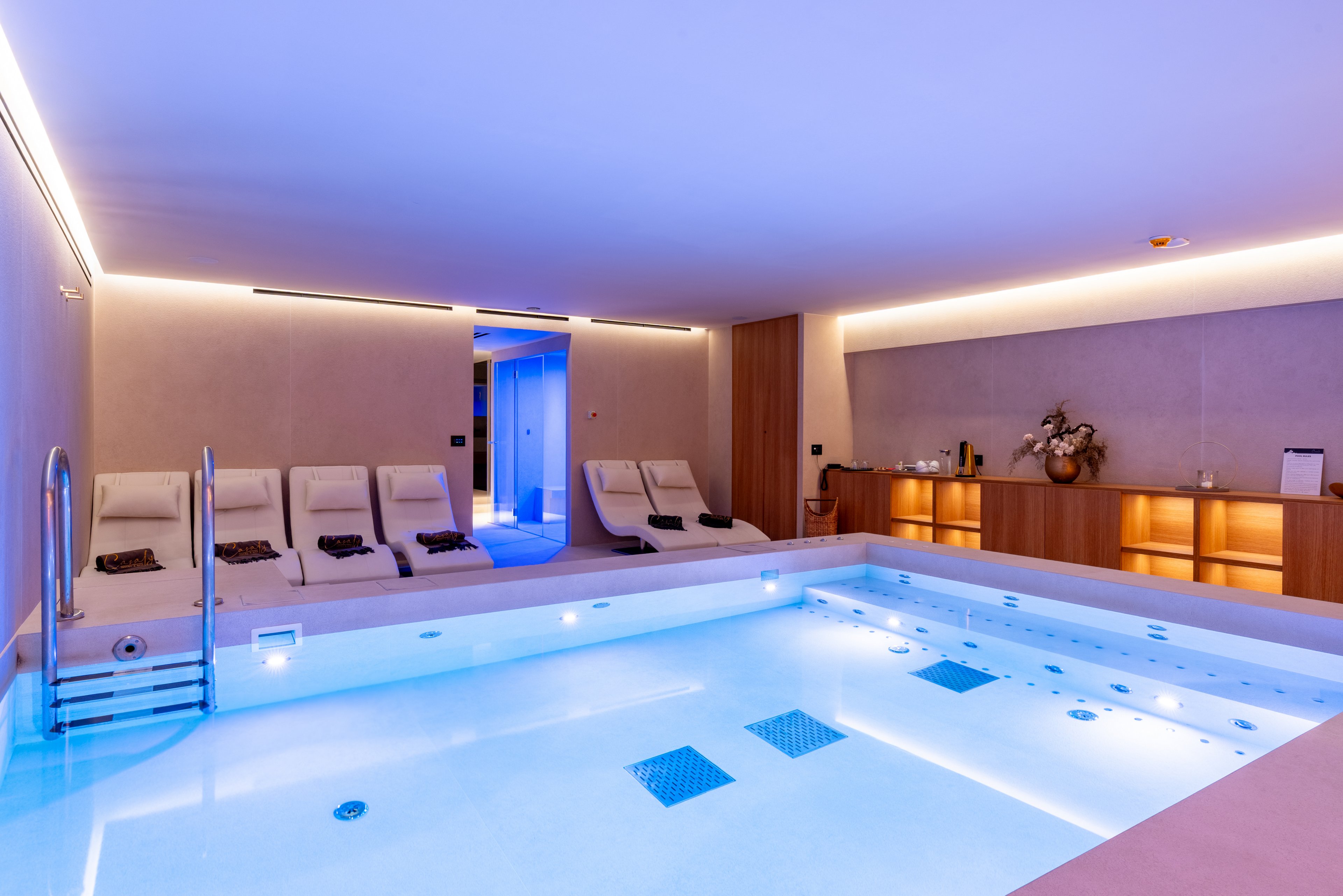 A contemporary indoor spa room features a large illuminated pool with built-in jets and underwater lights. White lounge chairs line one side of the space, each prepared with neatly folded dark towels. Warm wooden cabinetry and soft indirect lighting create a relaxing atmosphere. The overall design is minimalist and luxurious, emphasizing comfort and tranquility.