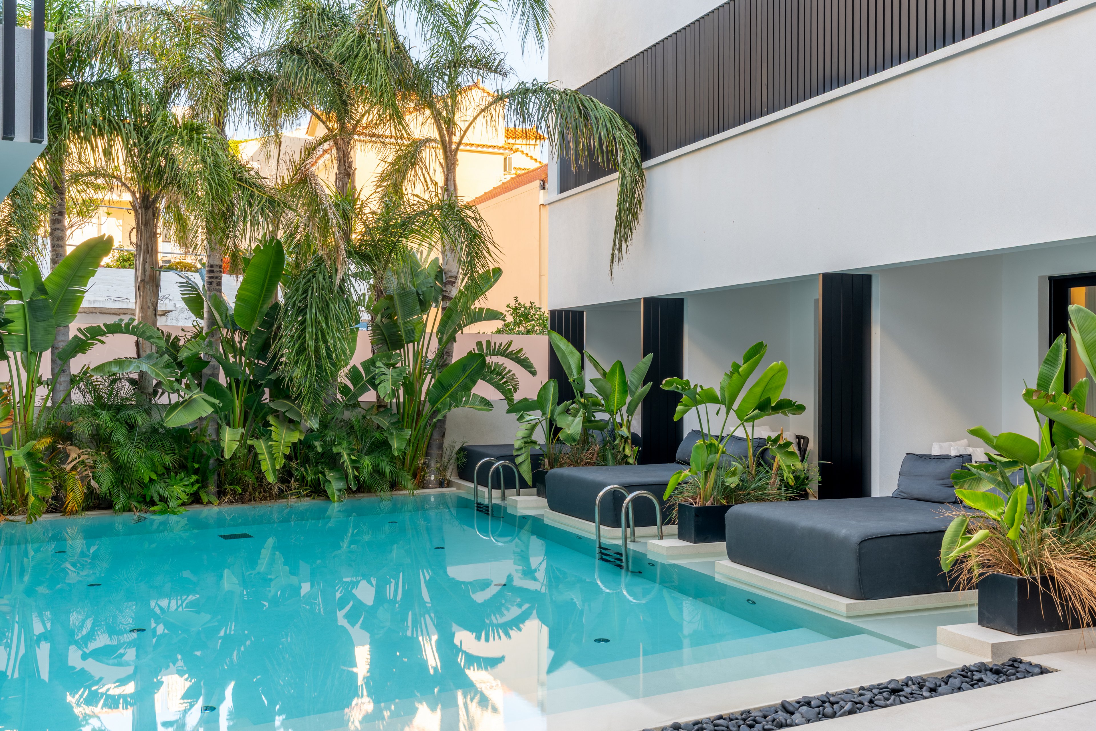 A contemporary outdoor swimming pool is surrounded by lush tropical plants and palm trees. Minimalist gray sun loungers sit partially over the water beside stainless steel pool ladders. The white building facade features clean lines and dark vertical accents, creating a stylish resort atmosphere. Soft reflections on the clear blue water enhance the calm, relaxing mood of the scene.