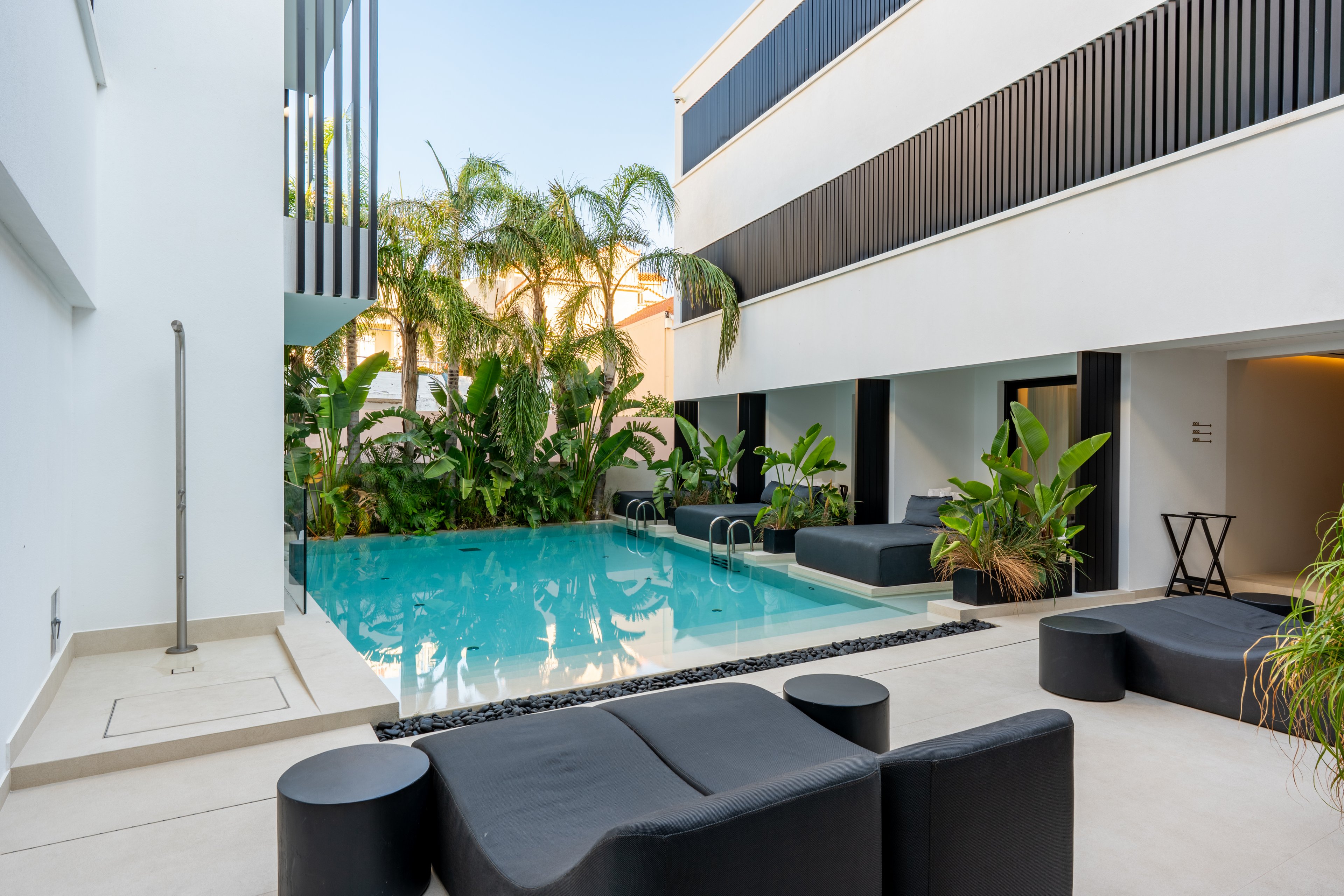 A contemporary courtyard features a rectangular swimming pool surrounded by lush tropical plants. Minimalist black lounge furniture and seating line the light-colored patio, creating a stylish relaxation area. Clean architectural lines and white walls frame the space, while dark horizontal accents add contrast. The tranquil water and greenery evoke a calm, resort-like atmosphere.
