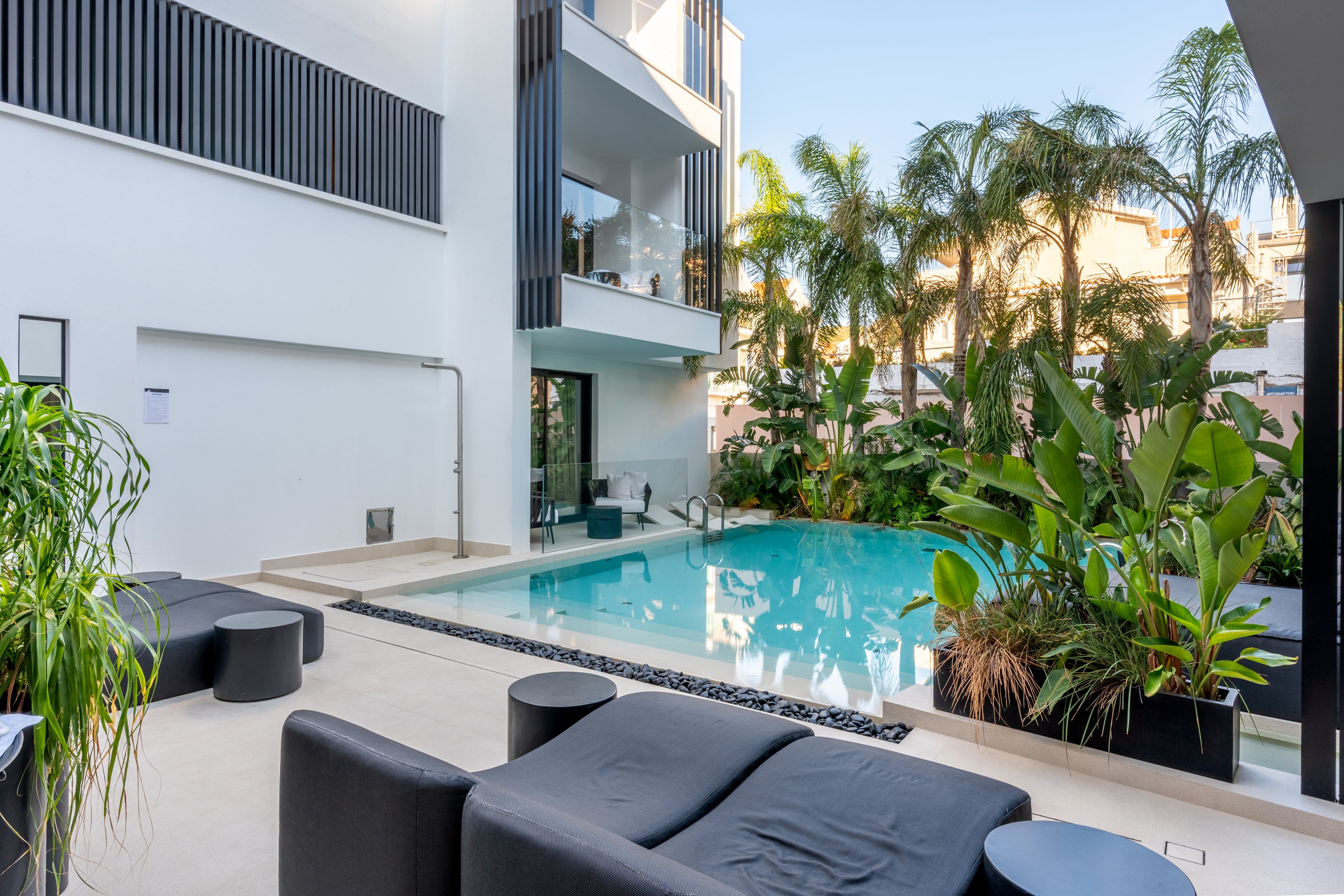 A contemporary residential courtyard features a clear blue swimming pool surrounded by tropical plants and palm trees. Minimalist black outdoor furniture sits on a light stone patio near the water. The white multi-story building has balconies and large glass windows overlooking the pool area. The scene is calm and inviting, suggesting a relaxed urban resort atmosphere.