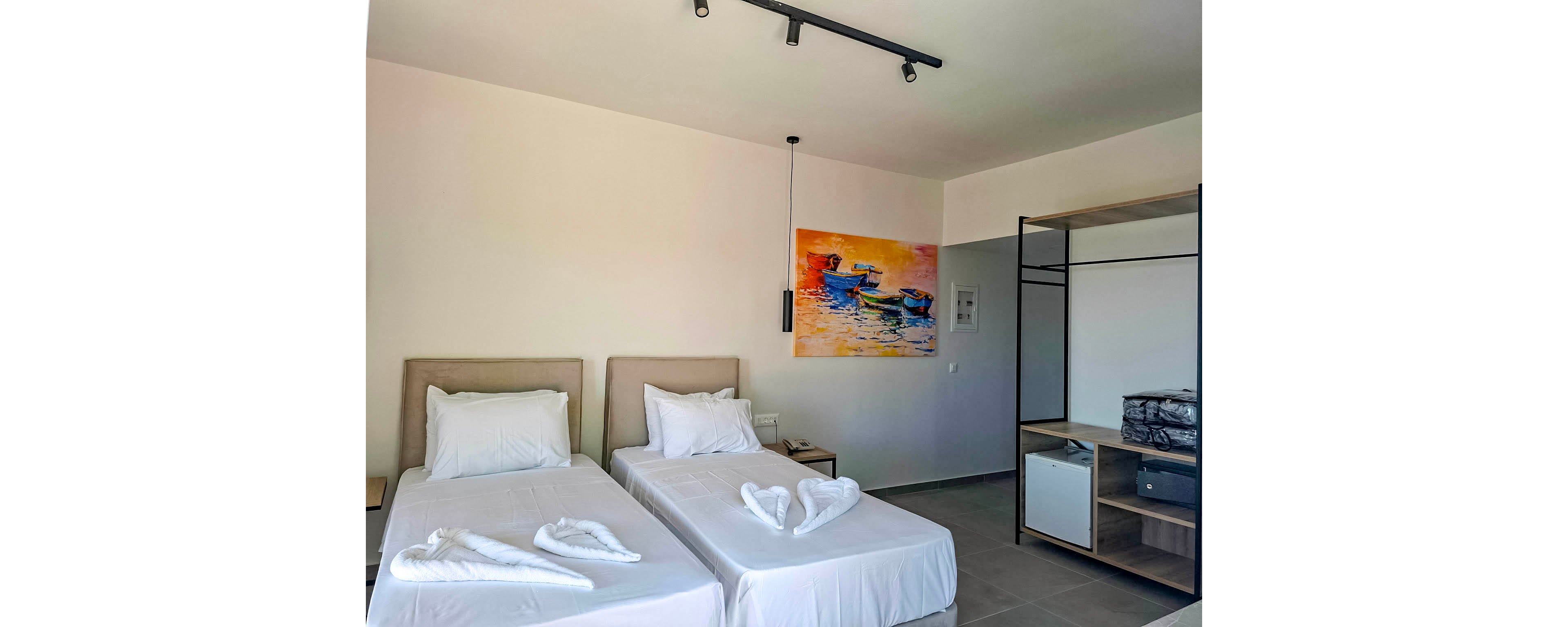 The image shows a contemporary hotel room featuring two single beds with crisp white linens. Towels are folded into heart shapes on each bed, adding a welcoming touch. The room includes minimalist furniture, an open wardrobe, and a colorful painting of boats on the wall. The overall palette is neutral with pops of color from the artwork, and the setting appears clean and organized.