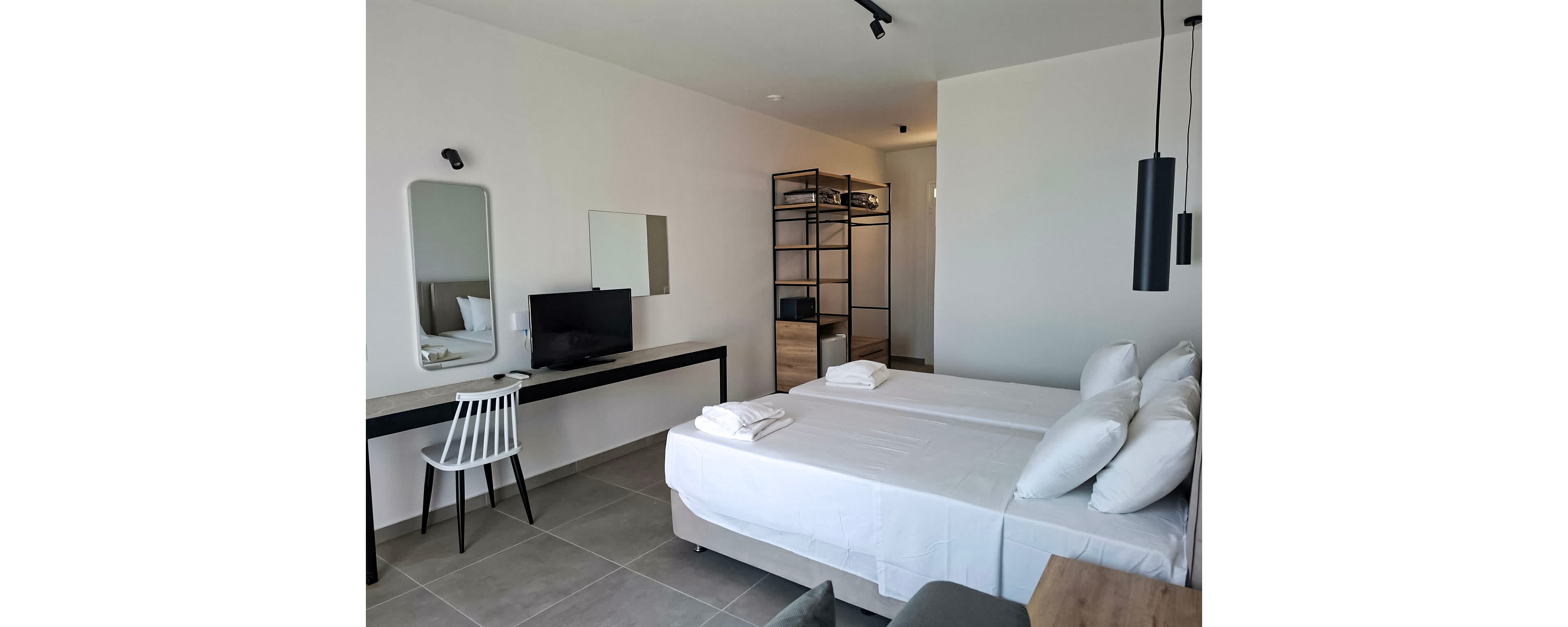A contemporary hotel room features two neatly made twin beds with white linens and towels. The space includes a sleek desk with a white chair, a wall-mounted mirror, and a flat-screen television. The open wardrobe and neutral color palette create a clean, minimalist atmosphere. Pendant lighting and tiled flooring add to the modern style.