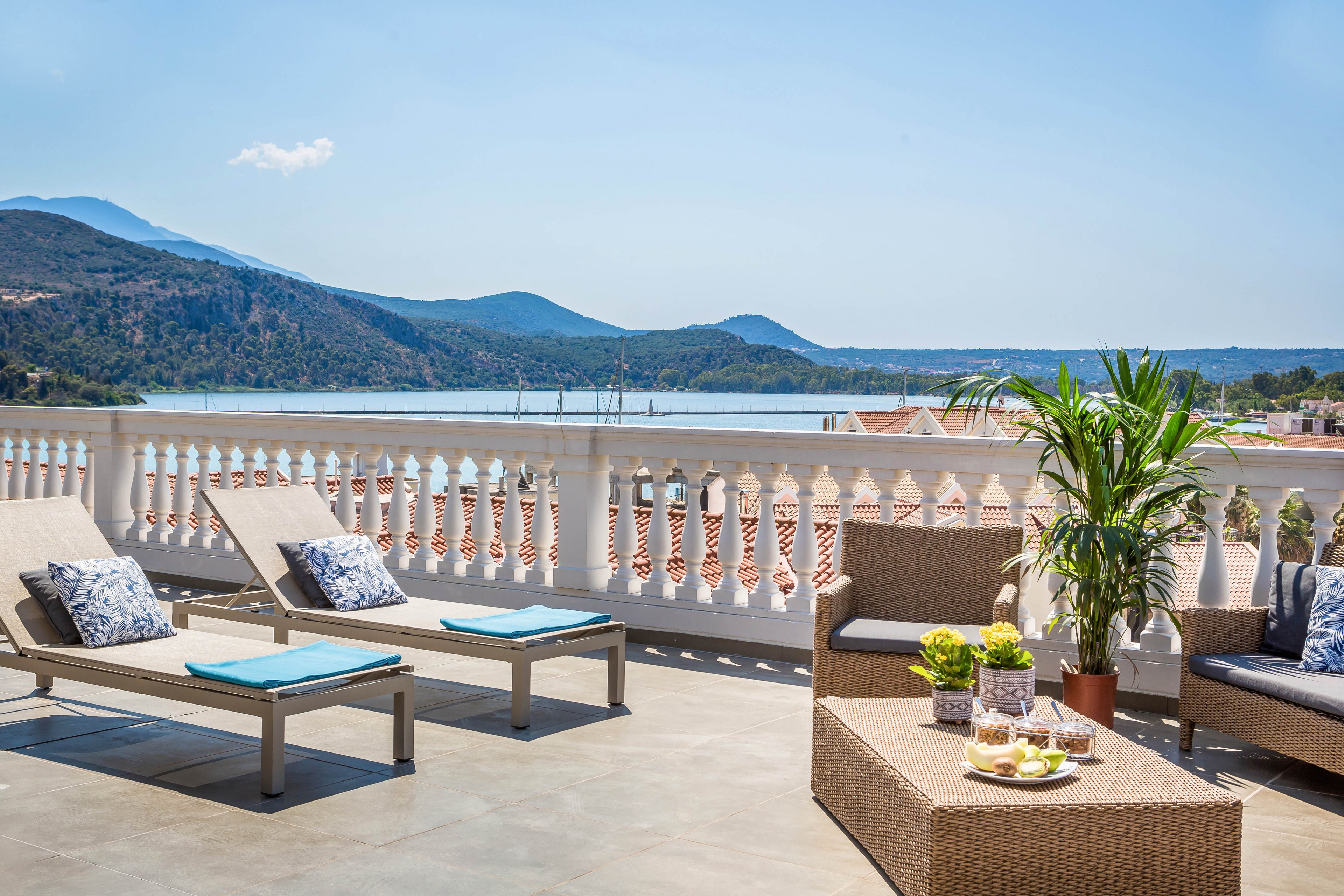 The main subject is a spacious outdoor terrace featuring wicker lounge chairs and a sofa, accented with decorative pillows and a coffee table set with refreshments. The setting overlooks a scenic mountain and lake landscape, framed by a white balustrade. The visuals highlight a bright, sunny atmosphere with clear skies, lush greenery, and a relaxed mood. No visible text or numeric values are present in the image.