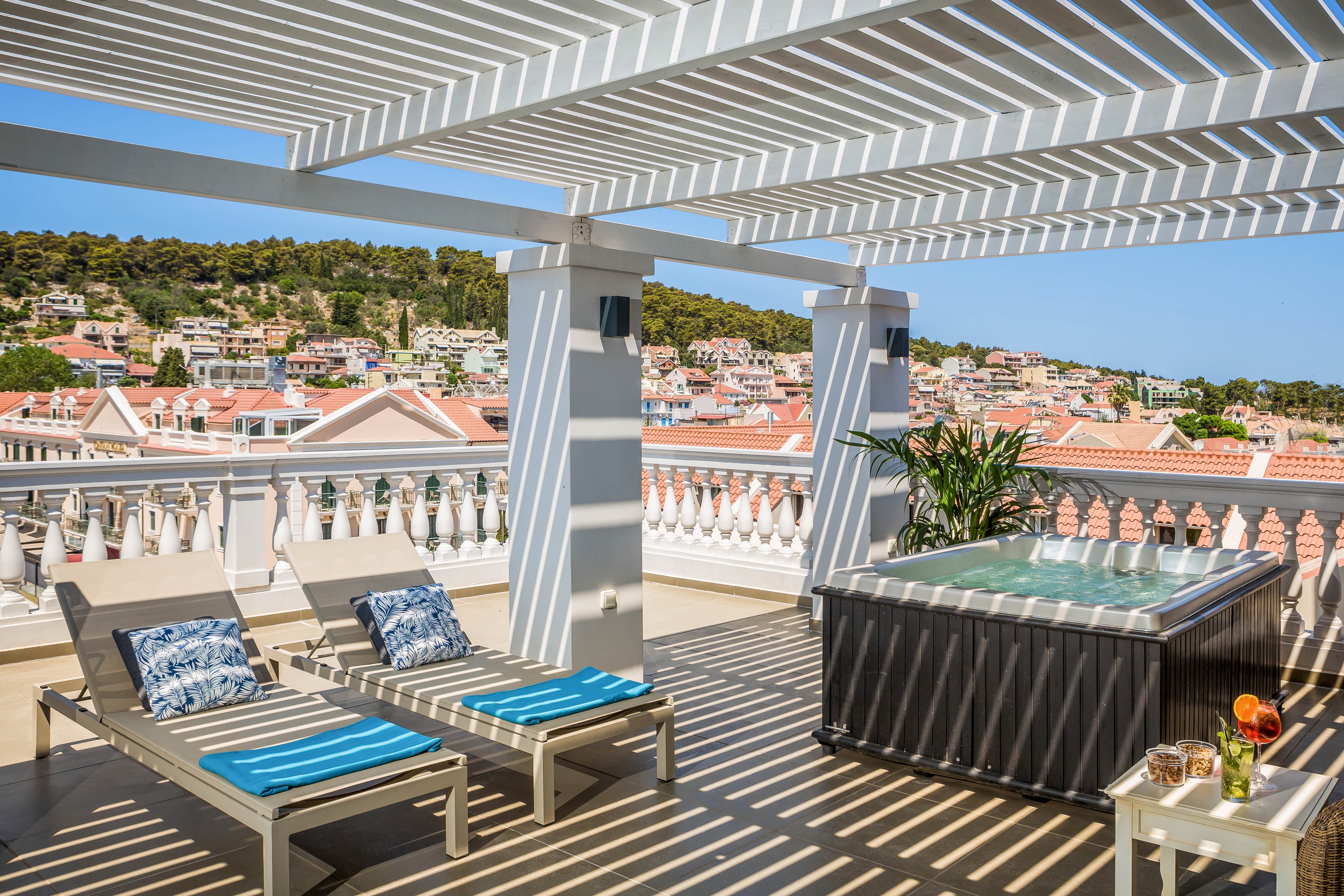 A spacious rooftop terrace features two cushioned lounge chairs and a modern hot tub under a white pergola. The setting overlooks a scenic hillside town with red-tiled rooftops and lush greenery. Sunlight casts striped shadows across the deck, creating a relaxing and inviting atmosphere. A small table holds drinks and fruit, enhancing the leisurely mood.