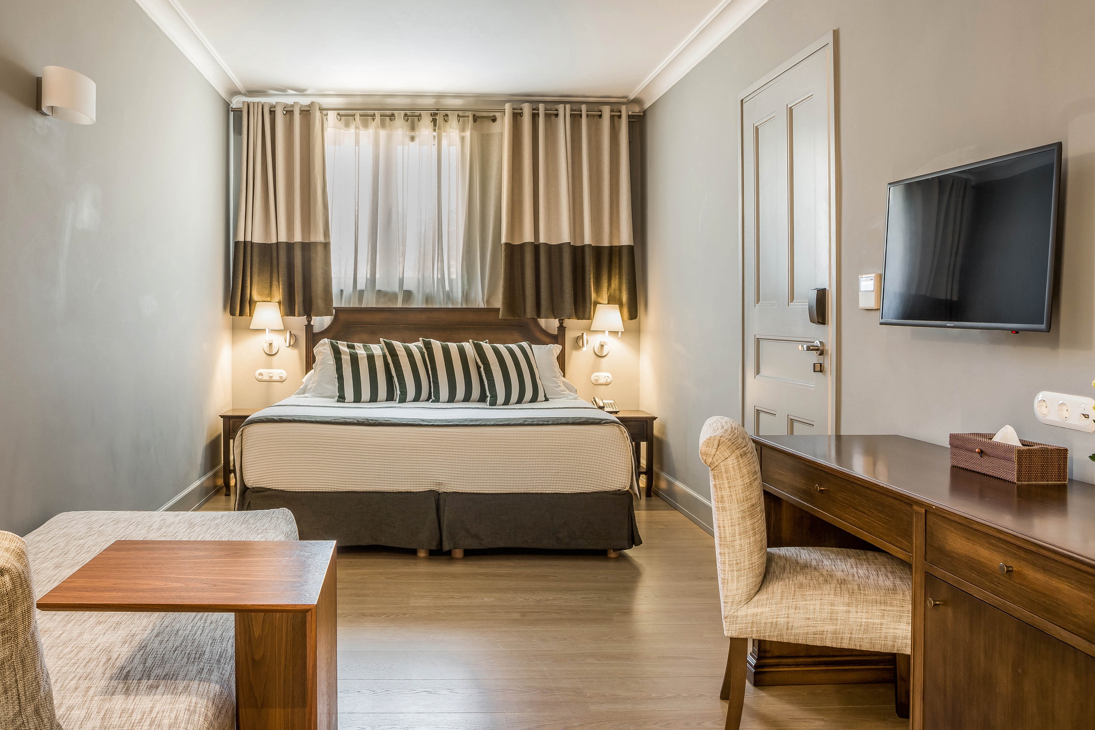 A contemporary hotel room features a neatly made double bed with striped pillows and matching curtains. The space includes a wooden desk, upholstered chair, and a wall-mounted flat-screen television. Soft lighting from bedside lamps and neutral tones create a calm, inviting atmosphere. The room is uncluttered, with a small seating area and a window providing natural light.