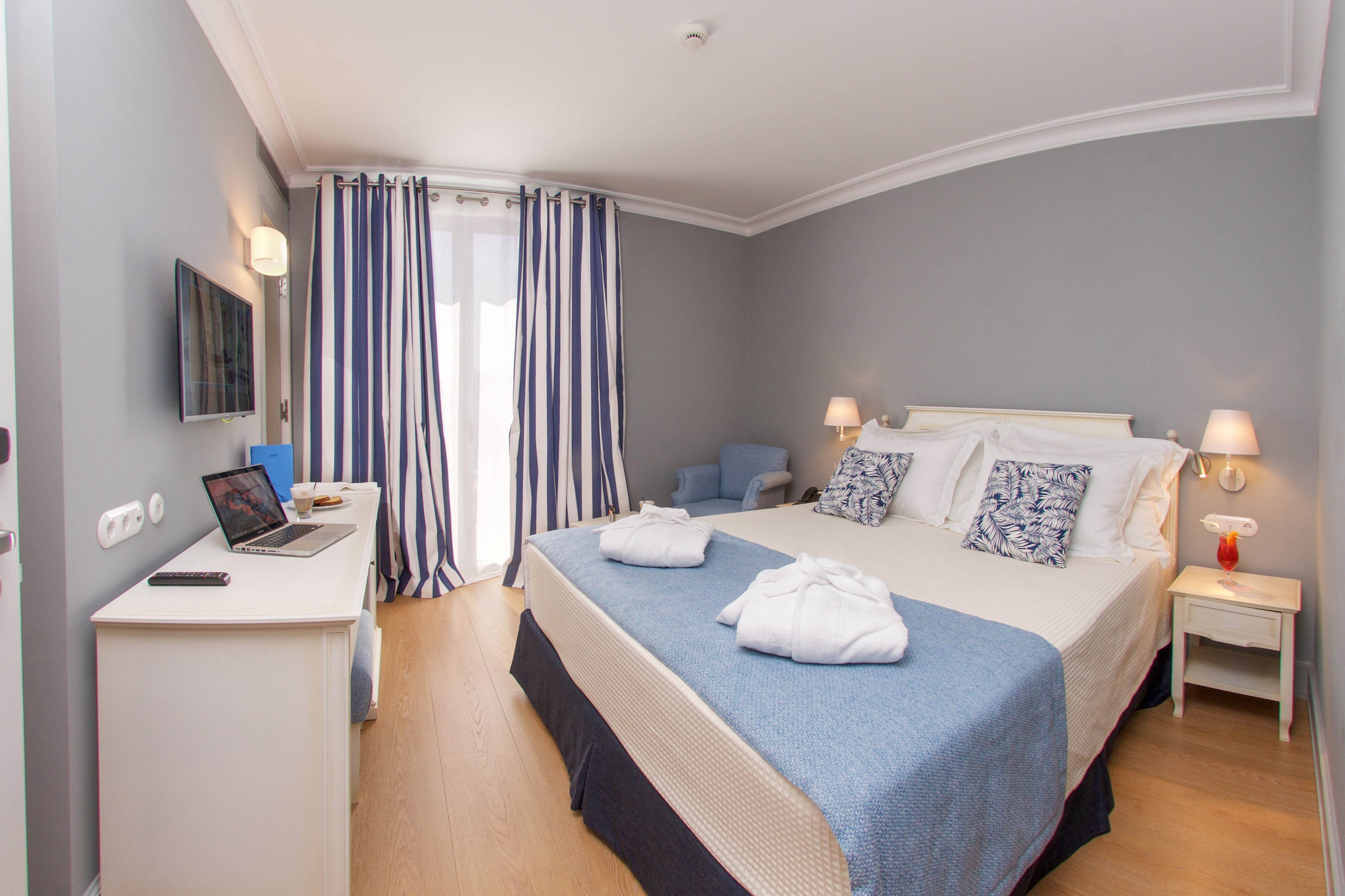 A neatly arranged hotel room features a double bed with blue and white bedding, two bathrobes, and decorative pillows. The space includes a desk with a laptop, a wall-mounted television, and striped curtains covering a window or balcony door. The room is well-lit with wall lamps and has a calm, inviting atmosphere with light wood flooring and soft blue accents.