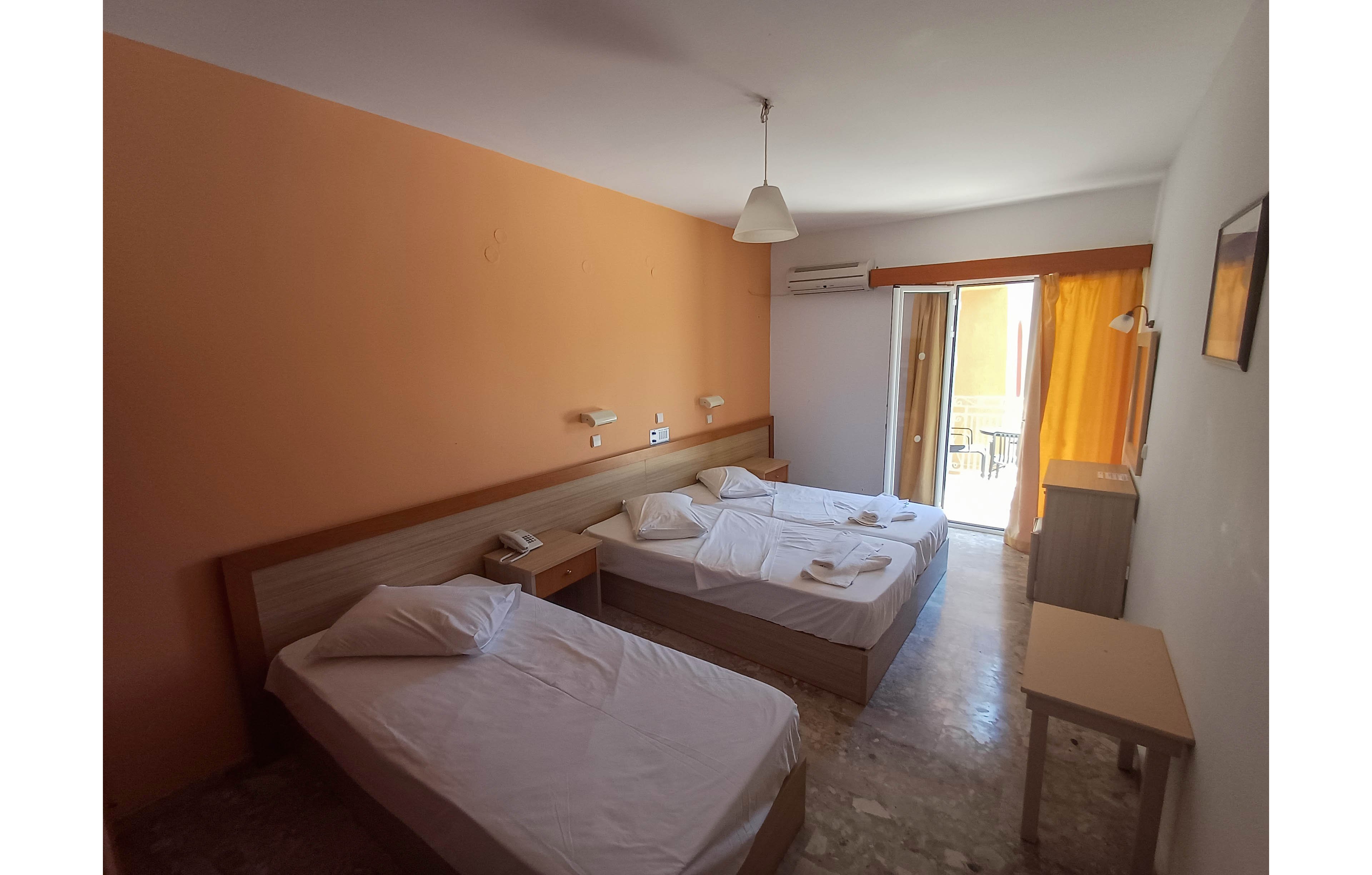 A modest hotel room features two single beds with white linens and folded towels placed on top. The space has light-colored walls, tiled flooring, and minimal furniture including a bedside table and a small desk. Natural light enters through a sliding glass door that opens to a balcony, complemented by a bright yellow curtain. An overhead pendant lamp and wall-mounted reading lights provide additional illumination, while an air conditioning unit is visible above the door.