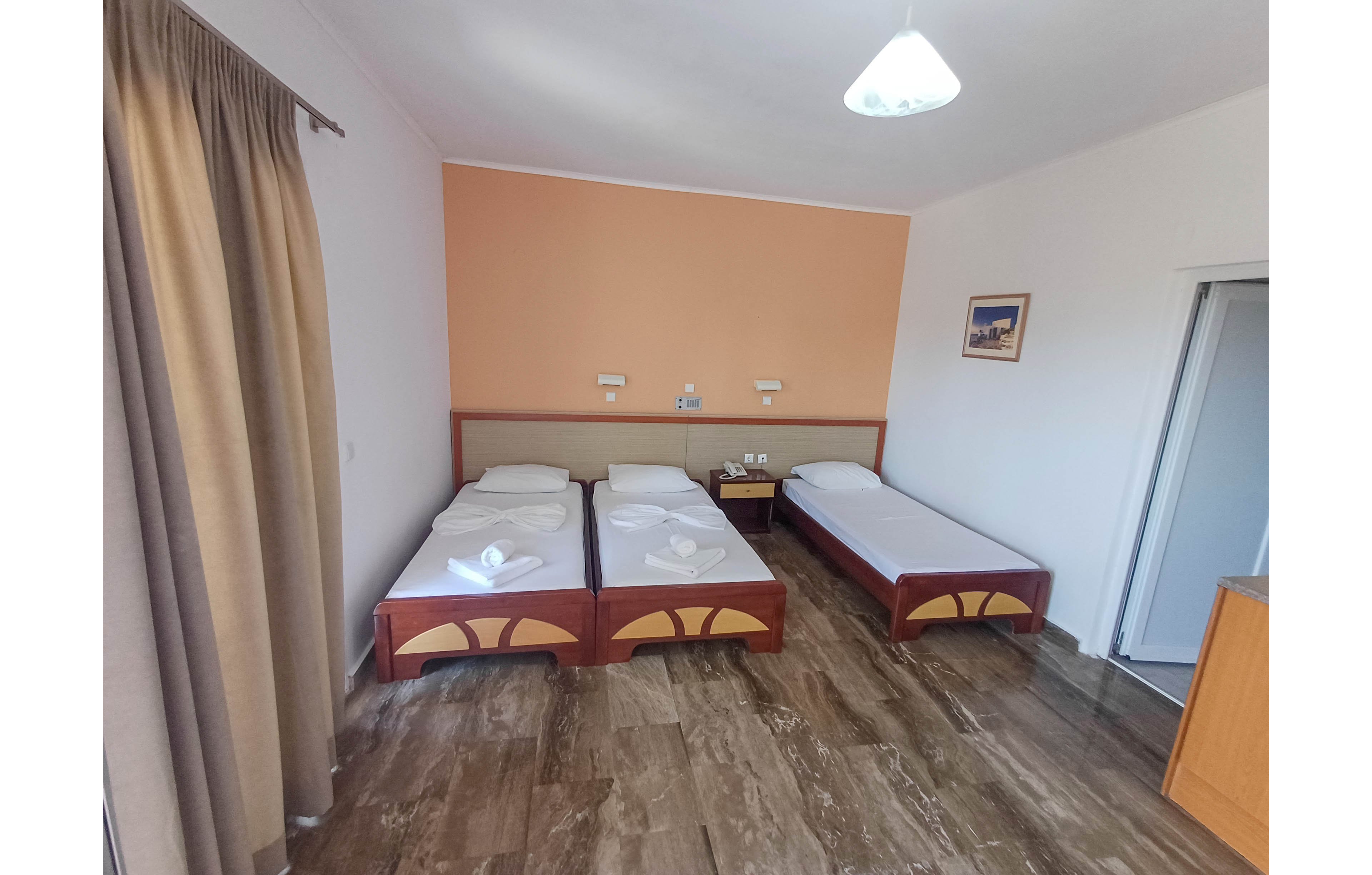 A simple hotel room features three single beds arranged along a peach-colored accent wall. Two beds are placed side by side with folded white towels, while a third bed stands separately near the corner. The room has tiled flooring, light walls, and a large curtain covering a window or balcony door. A small nightstand with outlets and switches, wall-mounted lights, and a framed picture complete the minimalist decor.