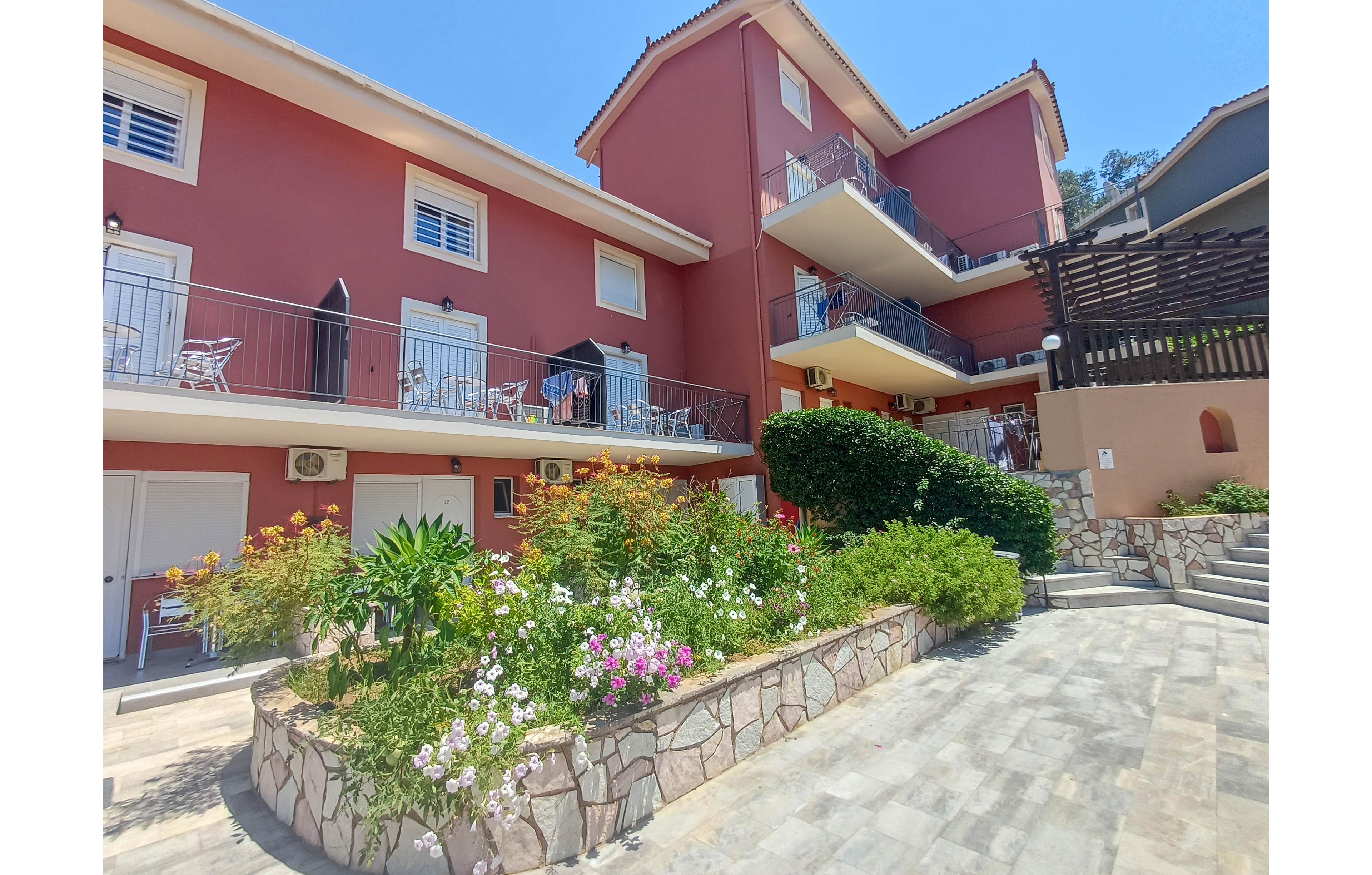 A multi-story red apartment building is shown with balconies overlooking a sunny courtyard. The foreground features a raised stone planter filled with colorful flowers and lush greenery. Outdoor furniture and railings line the balconies, suggesting a residential or vacation setting. Bright daylight and clear blue sky create a warm, inviting atmosphere.