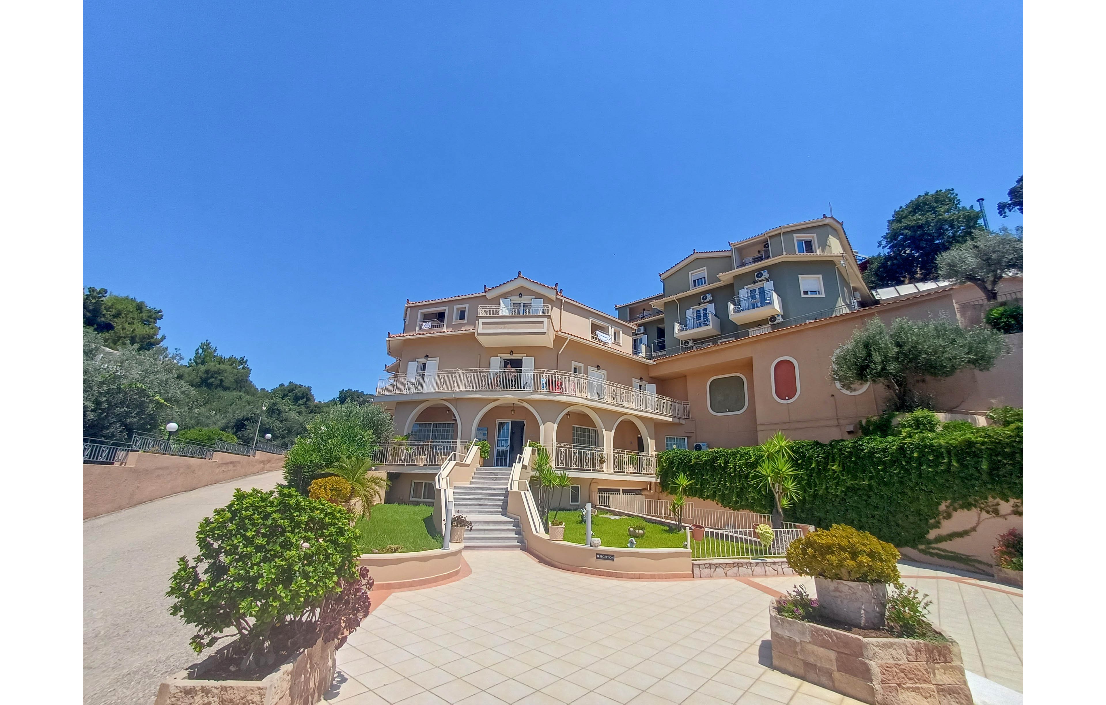 A multi-story Mediterranean-style villa complex stands prominently along a paved courtyard. The buildings feature arched balconies, light pastel facades, and white railings, surrounded by manicured shrubs and small palm trees. A central staircase leads up from the tiled walkway, emphasizing the grand entrance. The scene is bathed in bright daylight under a cloudless blue sky, creating a warm and inviting atmosphere.