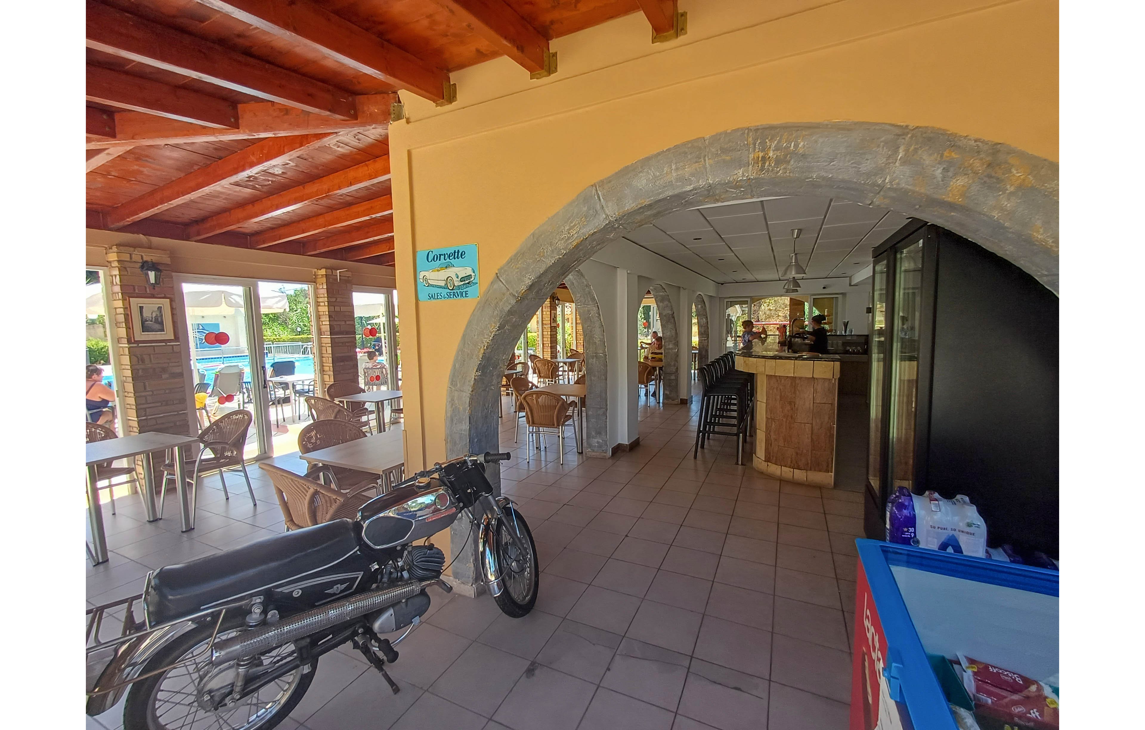 A classic motorbike is parked inside the tiled entrance of an open-air café. The space features arched stone doorways, wooden ceiling beams, and several tables and chairs arranged for guests. Bright daylight streams in from large windows, revealing greenery and a relaxed outdoor setting. A blue sign reading "Gerrette" hangs on the wall, and a counter area is visible deeper inside the café.