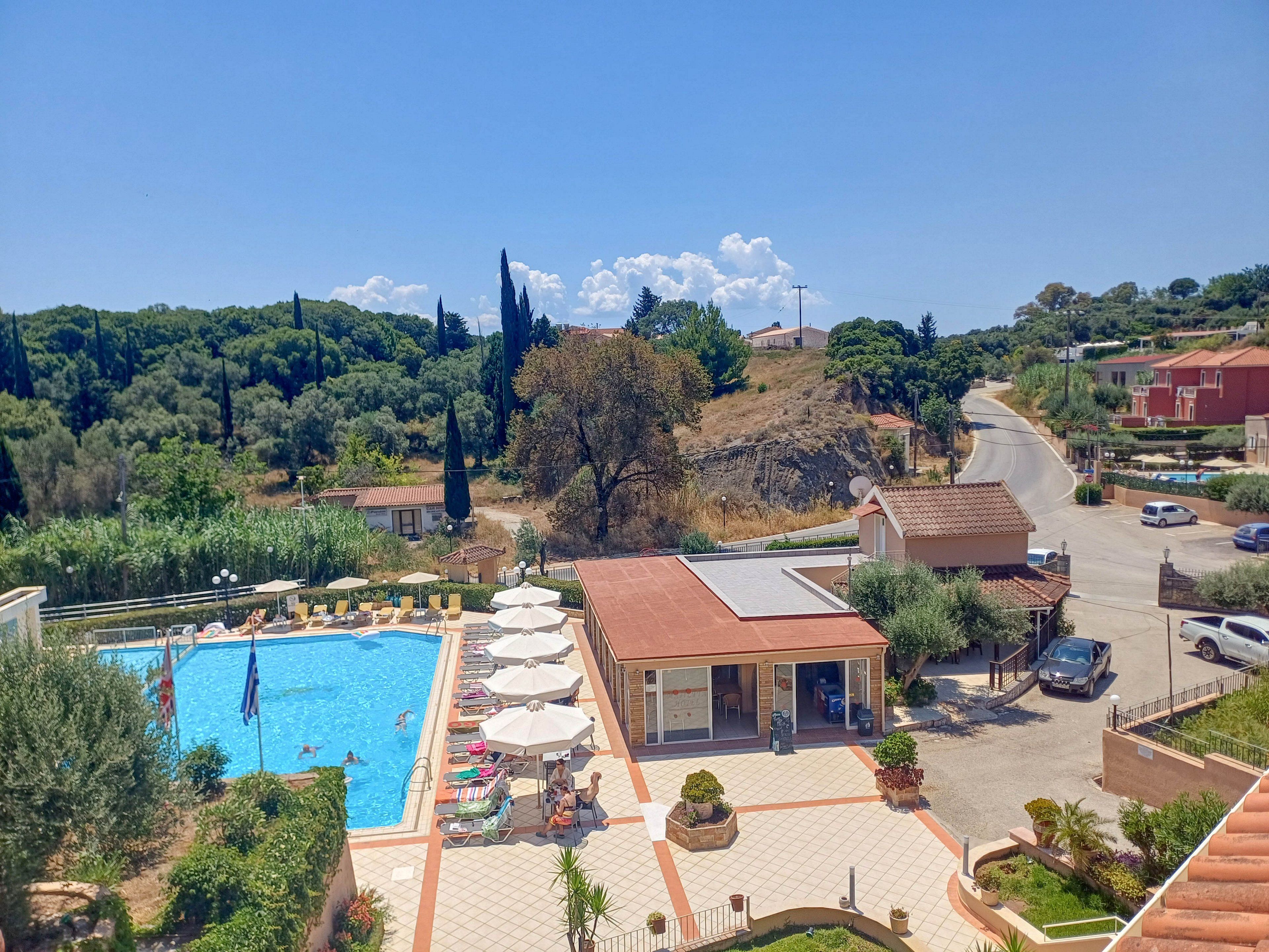 An outdoor swimming pool area is shown on a bright sunny day, surrounded by sun loungers and umbrellas. A small building and terrace sit next to the pool, with guests relaxing in the shade. Beyond the resort, a winding rural road passes by houses and parked cars. Lush green trees and hills form the backdrop under a clear blue sky with scattered clouds.