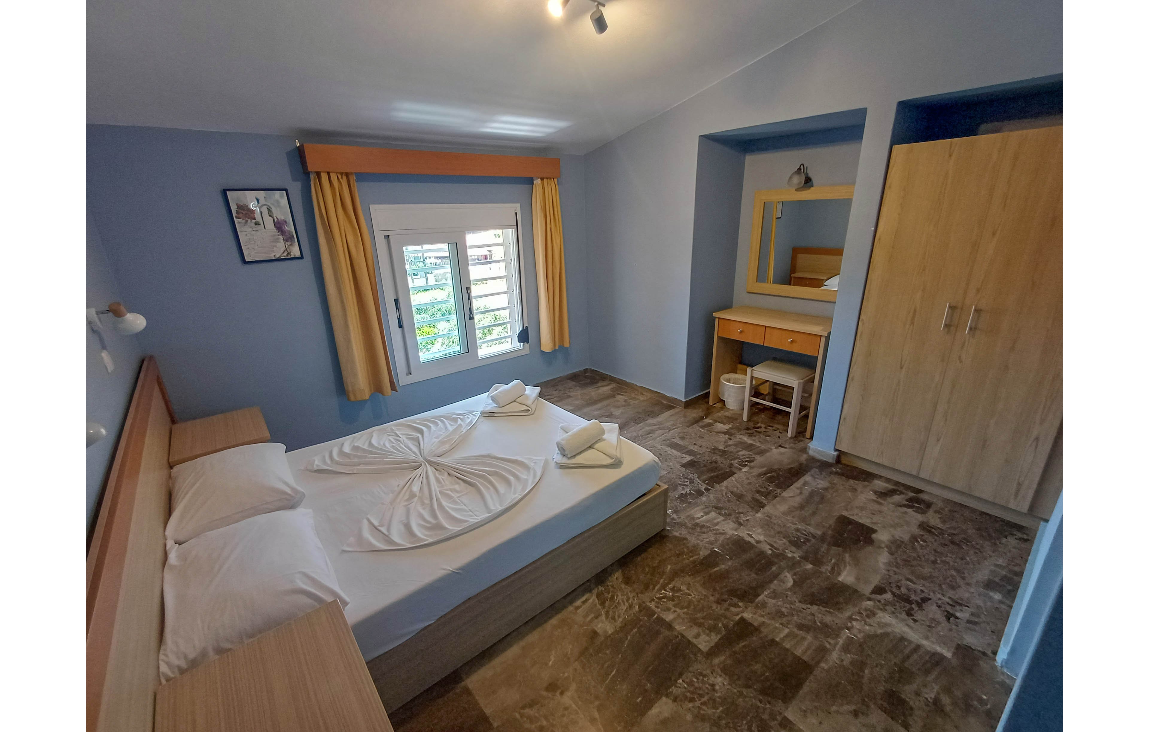 A neatly arranged double bed dominates a cozy hotel bedroom with light blue walls. Towels are folded decoratively on the bed, and pillows are stacked at the headboard. A window with yellow curtains lets in natural light and offers a view of greenery outside. On the right side, a wooden wardrobe and a dressing table with mirror and stool complete the simple, functional interior.
