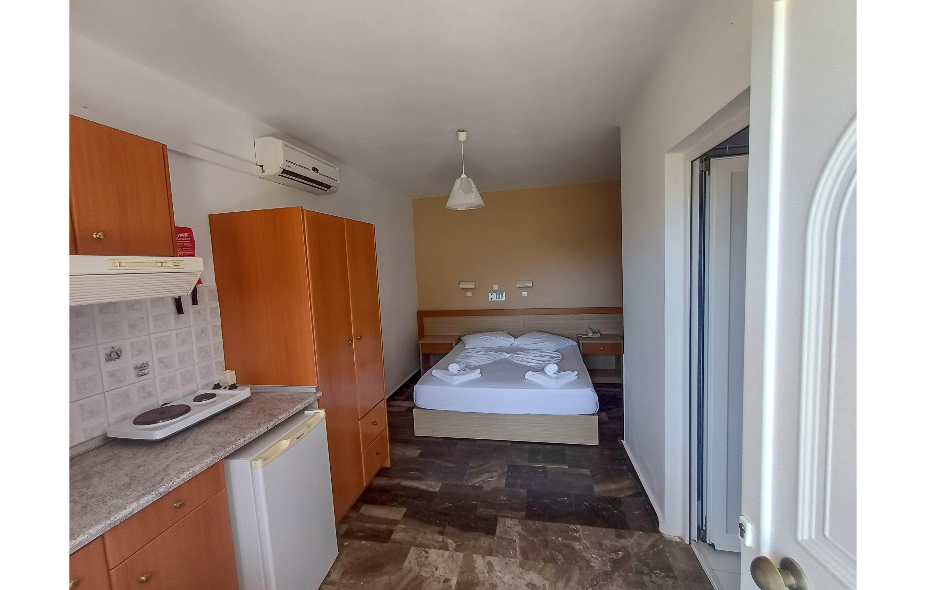 A small studio-style room features a double bed neatly made with white linens and towel swans. In the foreground, a compact kitchenette includes a countertop, electric cooktop, and mini refrigerator with wooden cabinets. The space has tiled flooring, a wall-mounted air conditioner, and neutral-colored walls. Natural light enters from a doorway on the right, giving the room a bright and simple appearance.