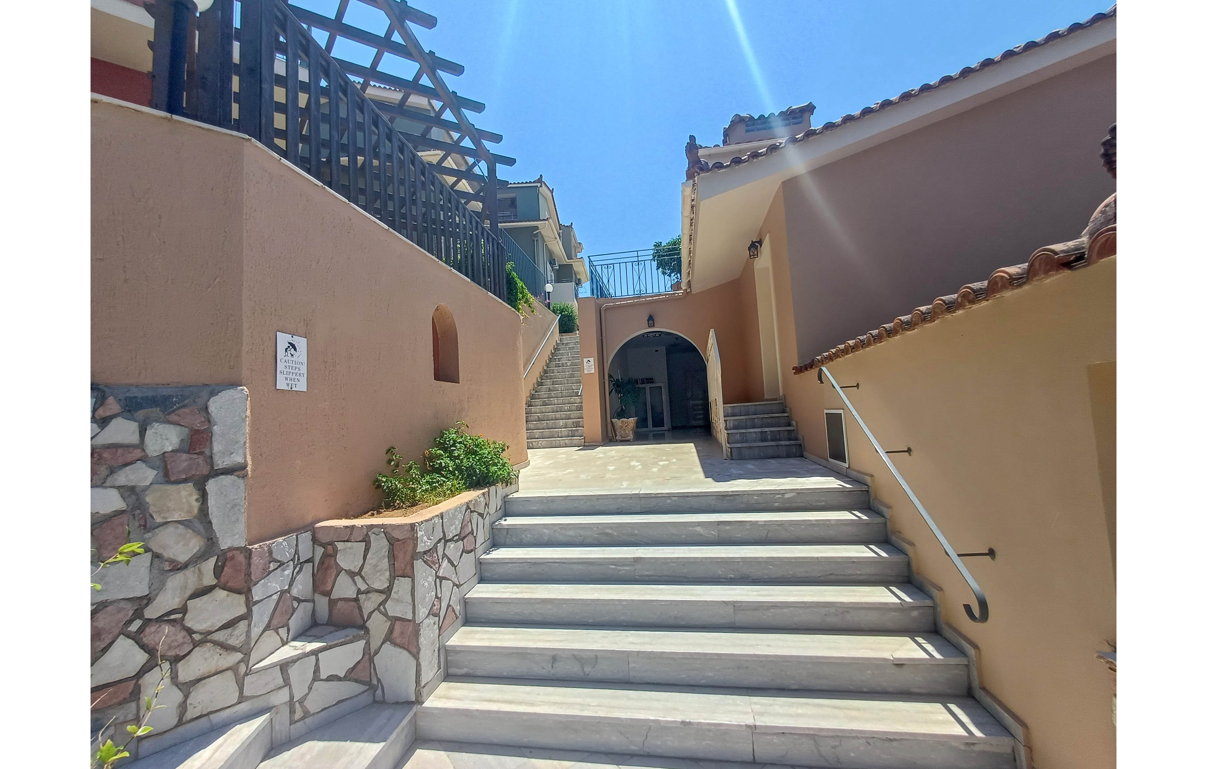 Outdoor marble stairs lead up between warm-toned stucco walls toward an arched doorway. The setting appears to be a Mediterranean-style villa or resort under bright midday sunlight. Decorative stonework lines the lower wall, and a metal handrail runs along the right side of the steps. A wooden pergola structure and tiled roofs add architectural interest against the clear blue sky.