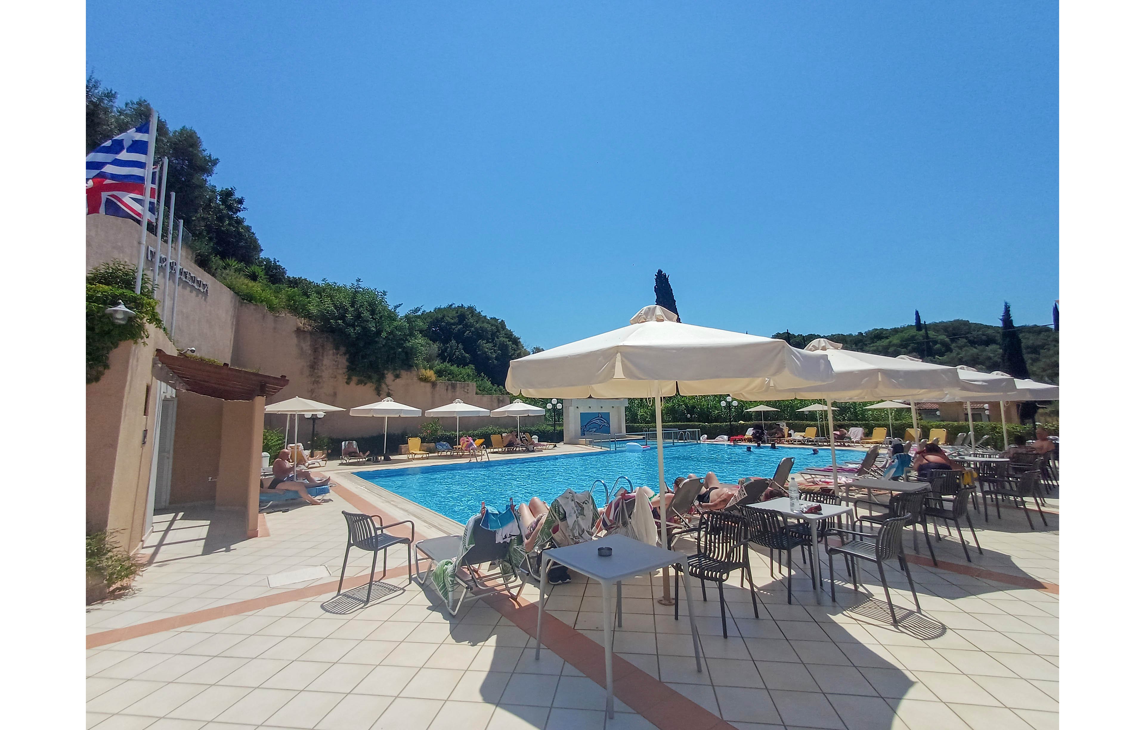 An outdoor resort swimming pool is surrounded by sun loungers and tables under large white umbrellas on a bright sunny day. Guests relax on chairs and recliners around the pool, enjoying the warm weather. The tiled terrace area leads to the water, with greenery and trees visible on the hillside in the background. Flags are raised on poles near the pool, adding a holiday atmosphere to the scene.