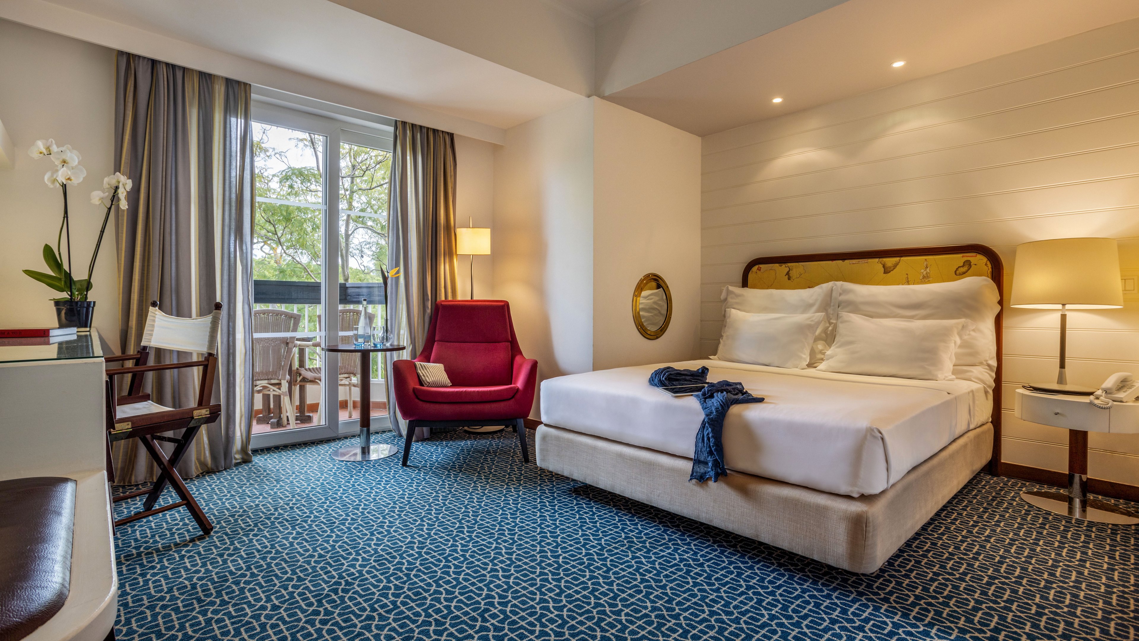 A neatly made double bed sits in a stylish hotel room with contemporary furnishings. A red armchair and floor lamp create a cozy reading corner near the balcony doors. Natural light enters through large windows with curtains, revealing an outdoor seating area. Blue patterned carpet, white walls, and decorative orchids add a clean, inviting atmosphere.