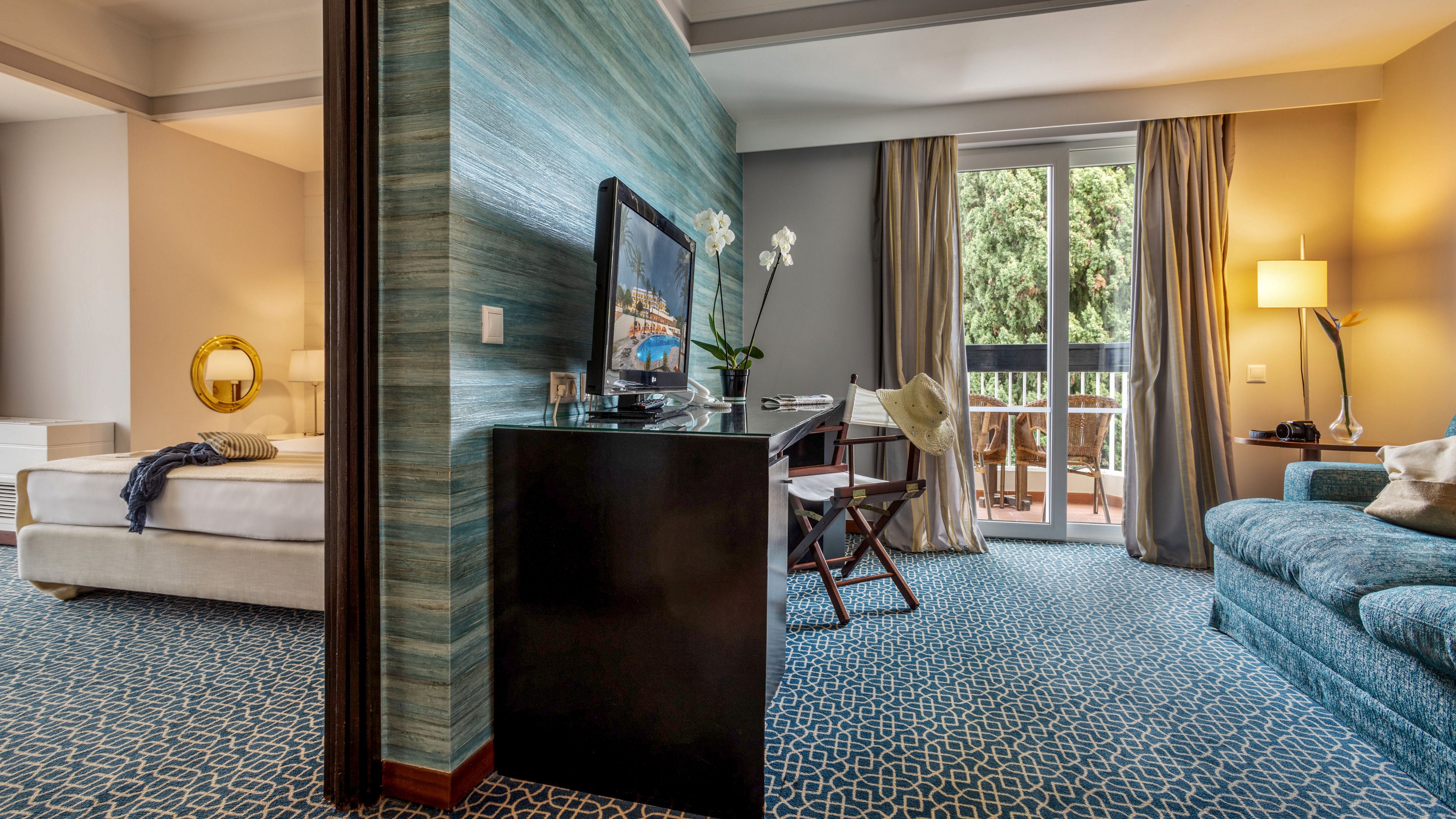 A contemporary hotel suite features a bedroom and living area separated by a partial wall. The living space includes a patterned blue carpet, teal sofa, and a desk with a flat-screen TV and orchid plant. Large glass doors open to a balcony with outdoor seating and a view of lush green trees. Warm lighting from floor and table lamps creates a cozy, inviting atmosphere.
