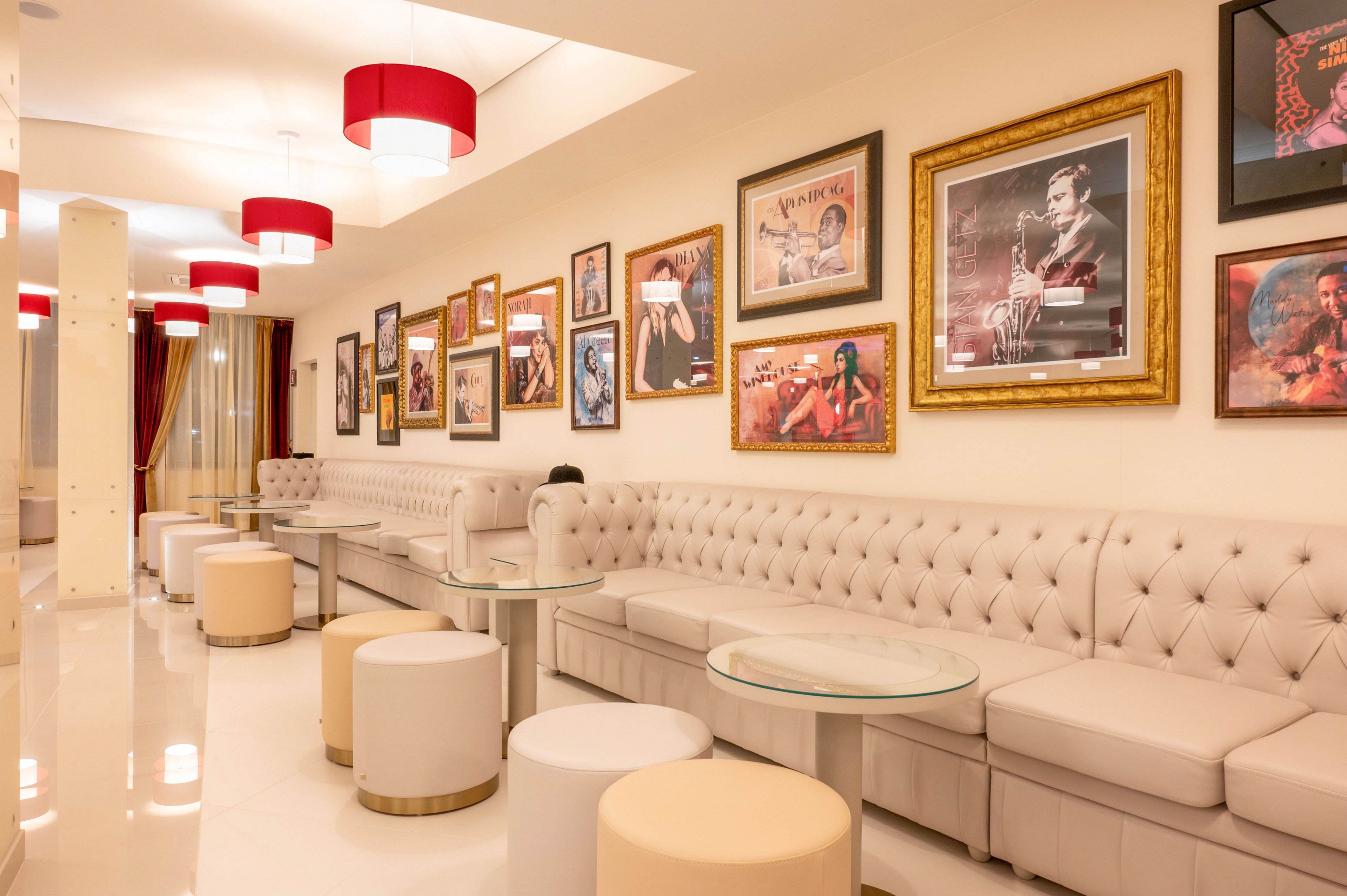 A modern lounge interior features a long tufted beige sofa lined with round ottomans and glass-topped tables. The walls are decorated with numerous framed artworks and posters, creating a gallery-like atmosphere. Red ceiling lamps and curtains add warm color accents to the otherwise light, neutral space. The polished floor and soft lighting contribute to a clean, inviting, and contemporary setting.