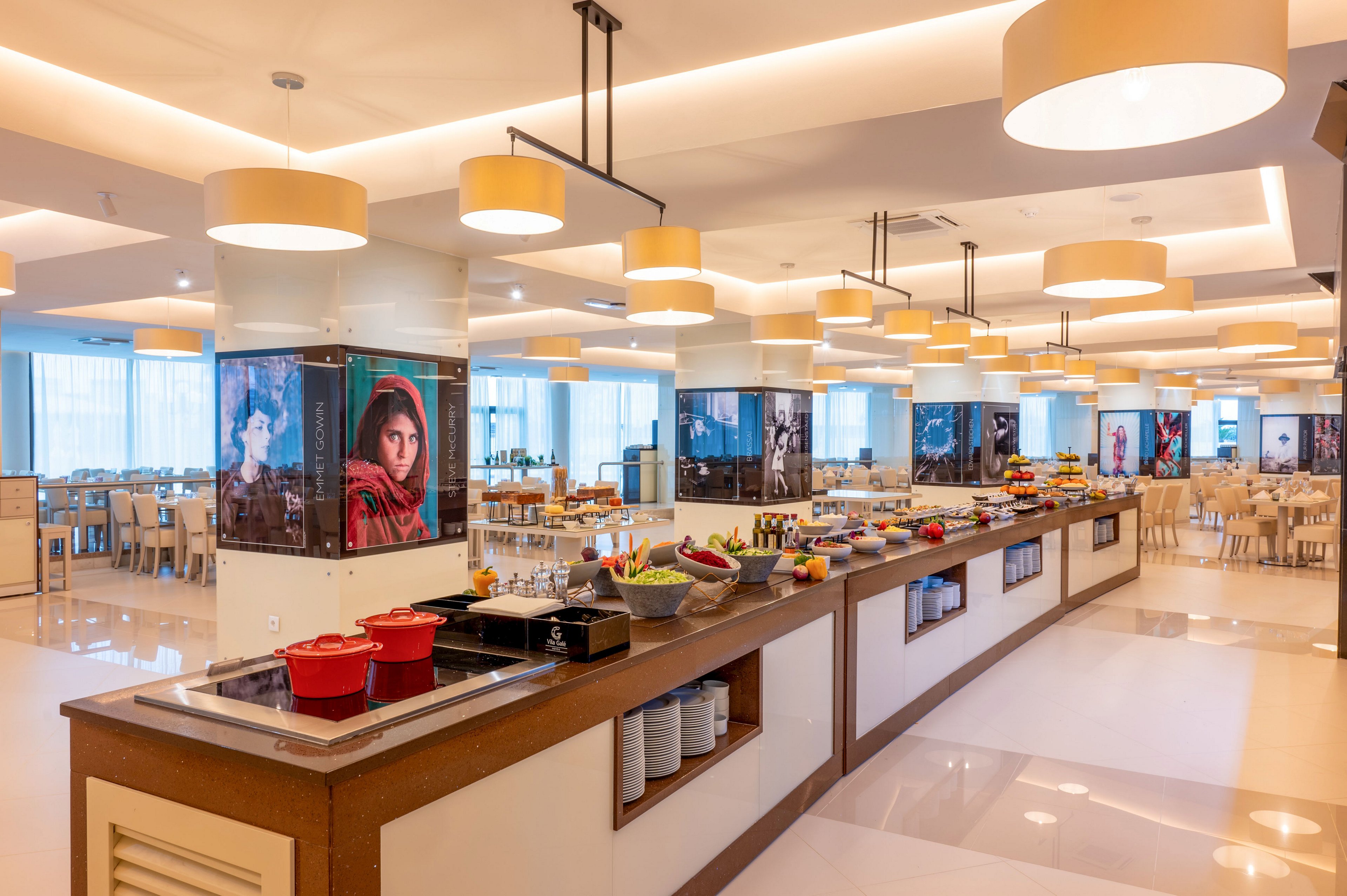 A spacious buffet-style restaurant features a long central serving island with various dishes and condiments arranged neatly. Warm ceiling lights and large windows create a bright, inviting atmosphere with reflections on the polished floor. Framed portraits decorate the columns, adding visual interest around the dining area. Numerous tables and chairs are set up in the background, ready to accommodate guests.