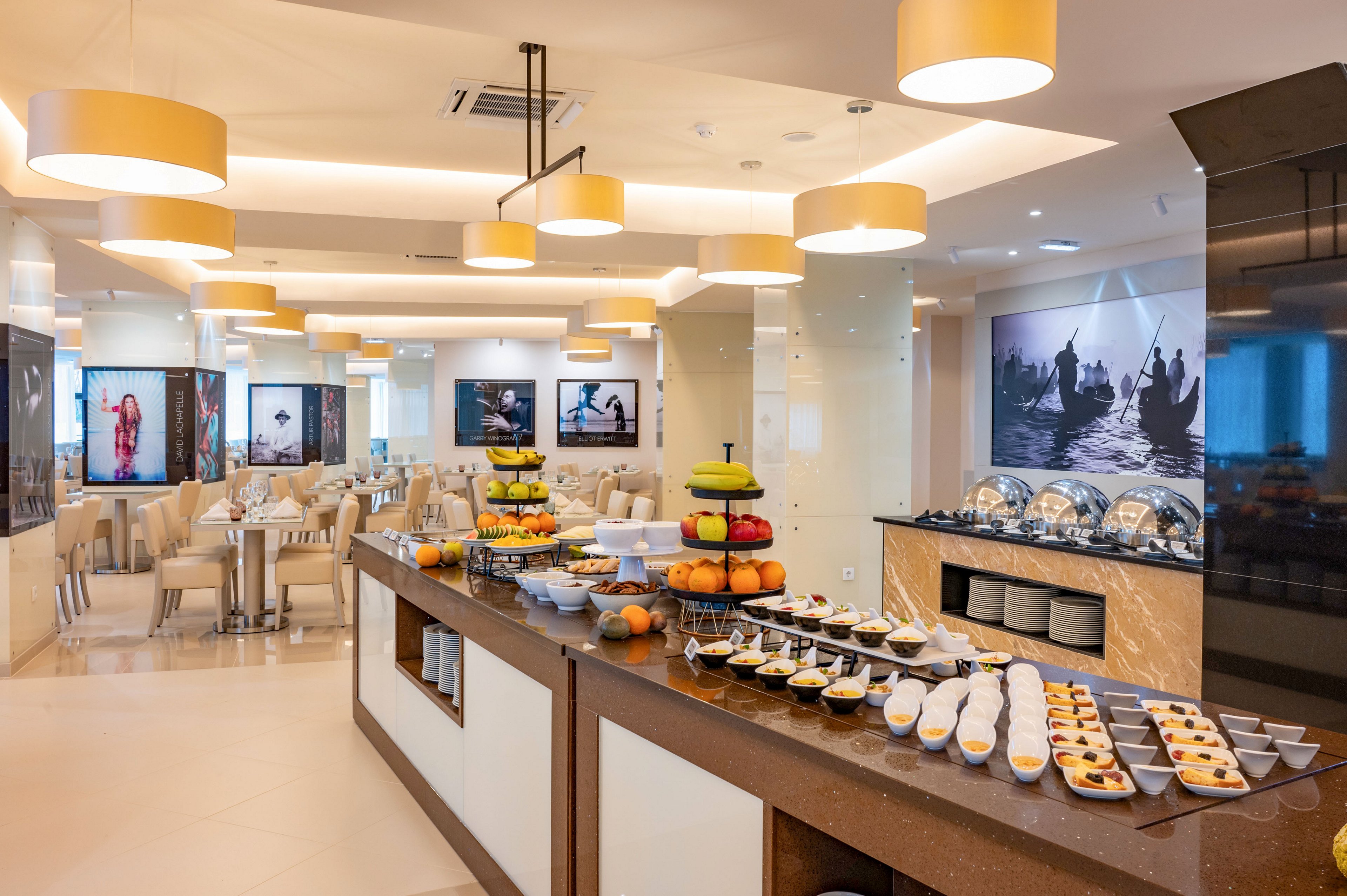 A spacious, modern dining room features a long buffet counter filled with desserts, pastries, and fresh fruit. Stainless steel chafing dishes line the back counter, suggesting a hot meal service in a hotel or restaurant setting. Warm pendant lights and neutral beige tones create a bright, inviting atmosphere. Large wall-mounted screens or artworks add a contemporary touch to the interior decor.