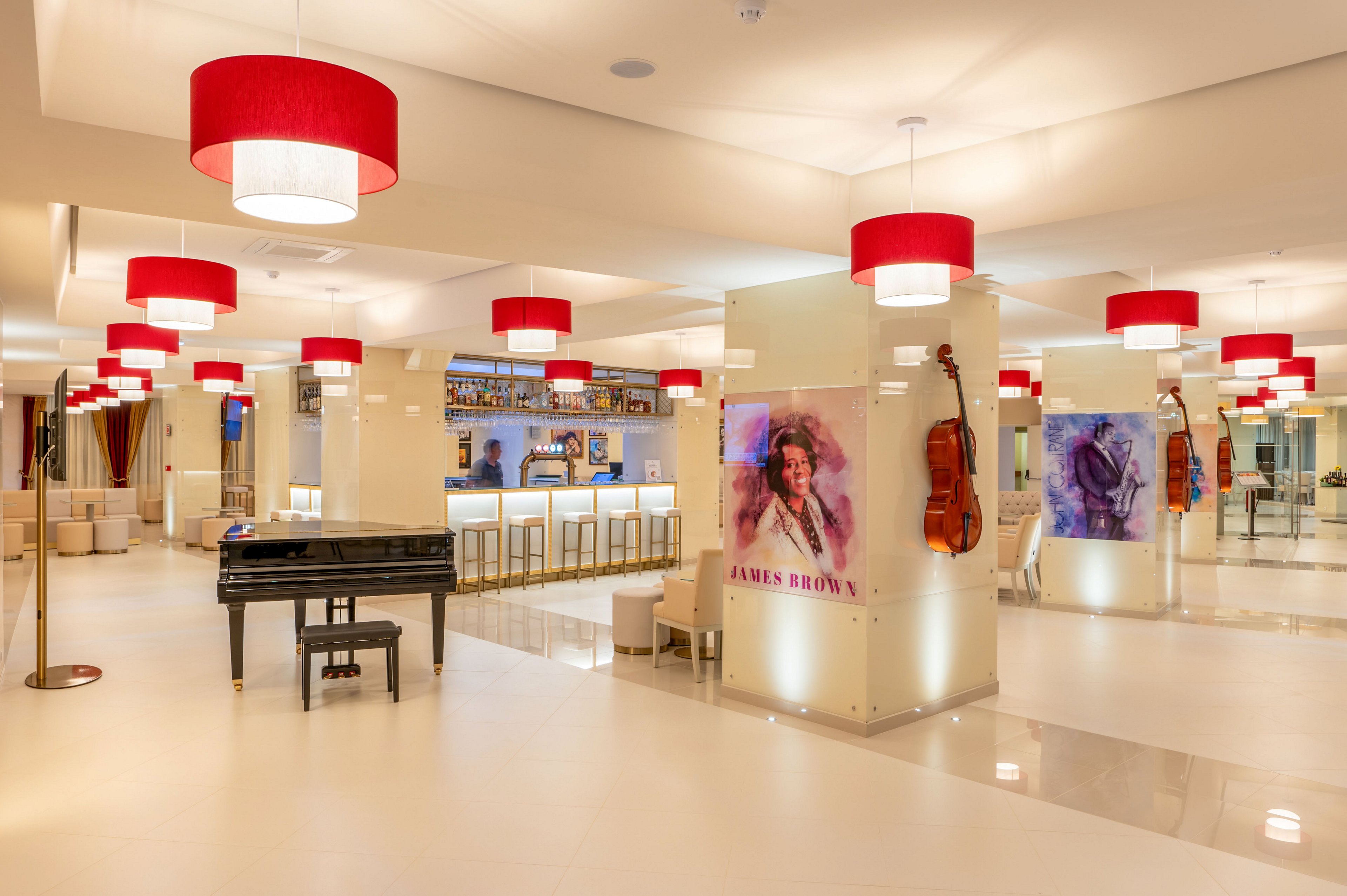 Bright contemporary lounge interior features a sleek bar area and a black grand piano in the foreground. Red pendant lights hang from the ceiling, contrasting with the neutral white floors and walls. Large artistic portraits of musicians and mounted violins decorate the columns, adding a musical theme to the space. Bar stools line the counter, creating a welcoming social atmosphere in this stylish hotel or venue lobby.