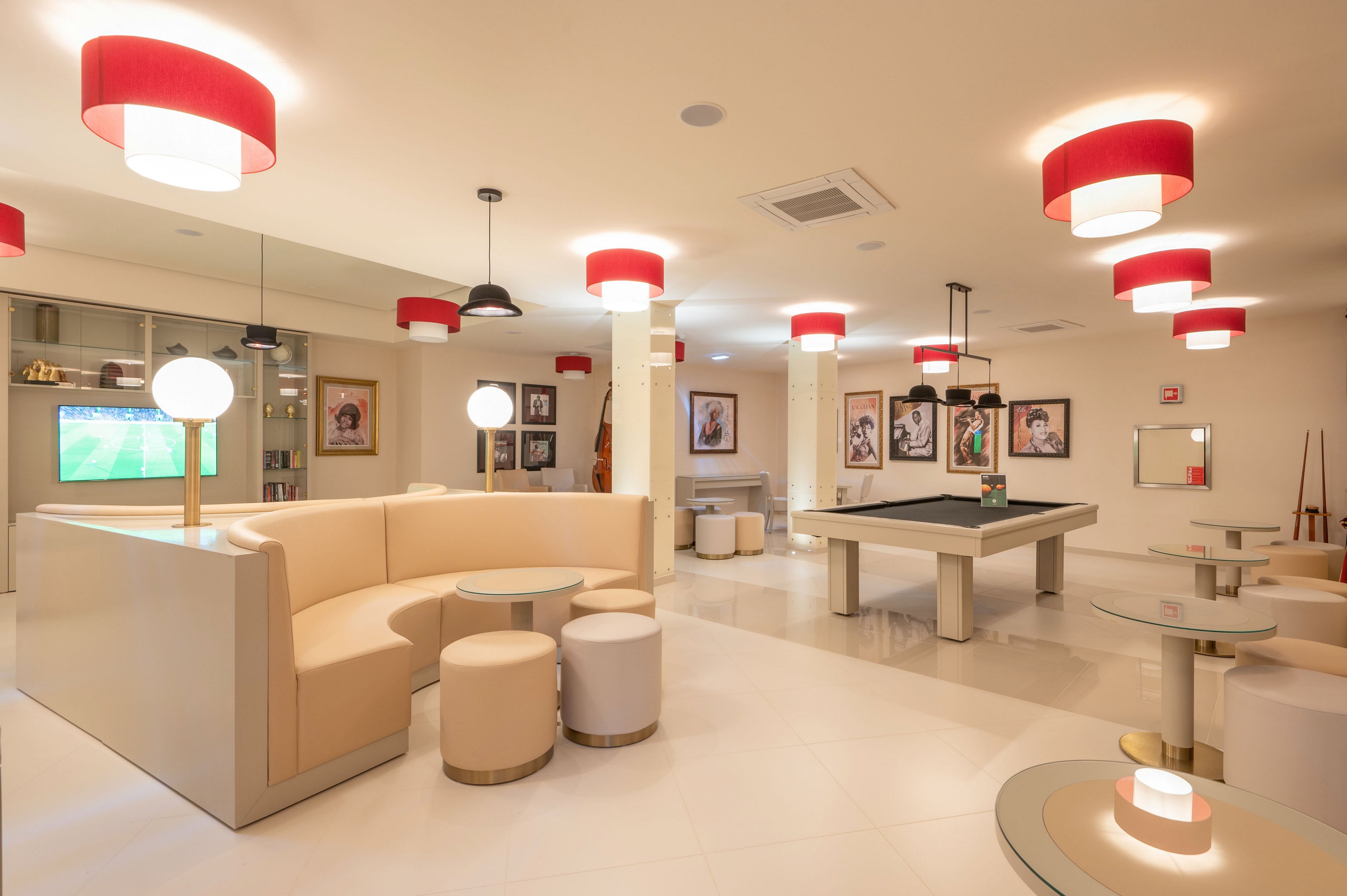 A contemporary lounge features a large beige sectional sofa and round stools arranged around sleek glass tables. A pool table stands at the center of the room, with cues and accessories visible nearby. Red and white ceiling lights create a warm, inviting glow over the polished floor. Framed artwork decorates the walls, and a television screen in the background shows a sports game, enhancing the entertainment atmosphere.