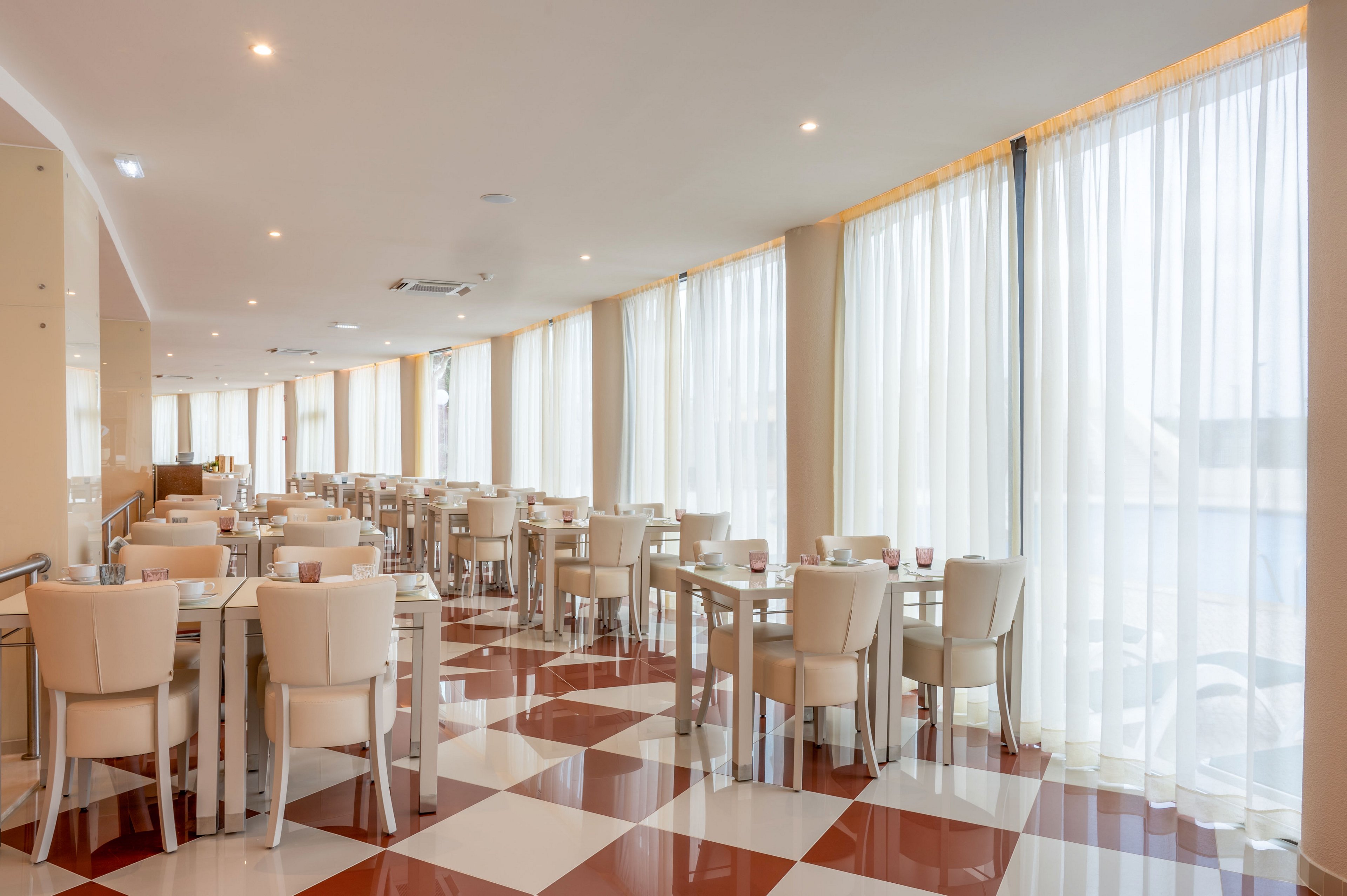 Spacious restaurant dining area with rows of tables and cream-colored chairs is shown in soft daylight. Large floor-to-ceiling windows with sheer white curtains line one side of the room, creating an airy atmosphere. The floor features a glossy red and white checkerboard pattern that adds visual interest. Table settings are minimal, suggesting a clean, contemporary style suitable for casual or formal dining.