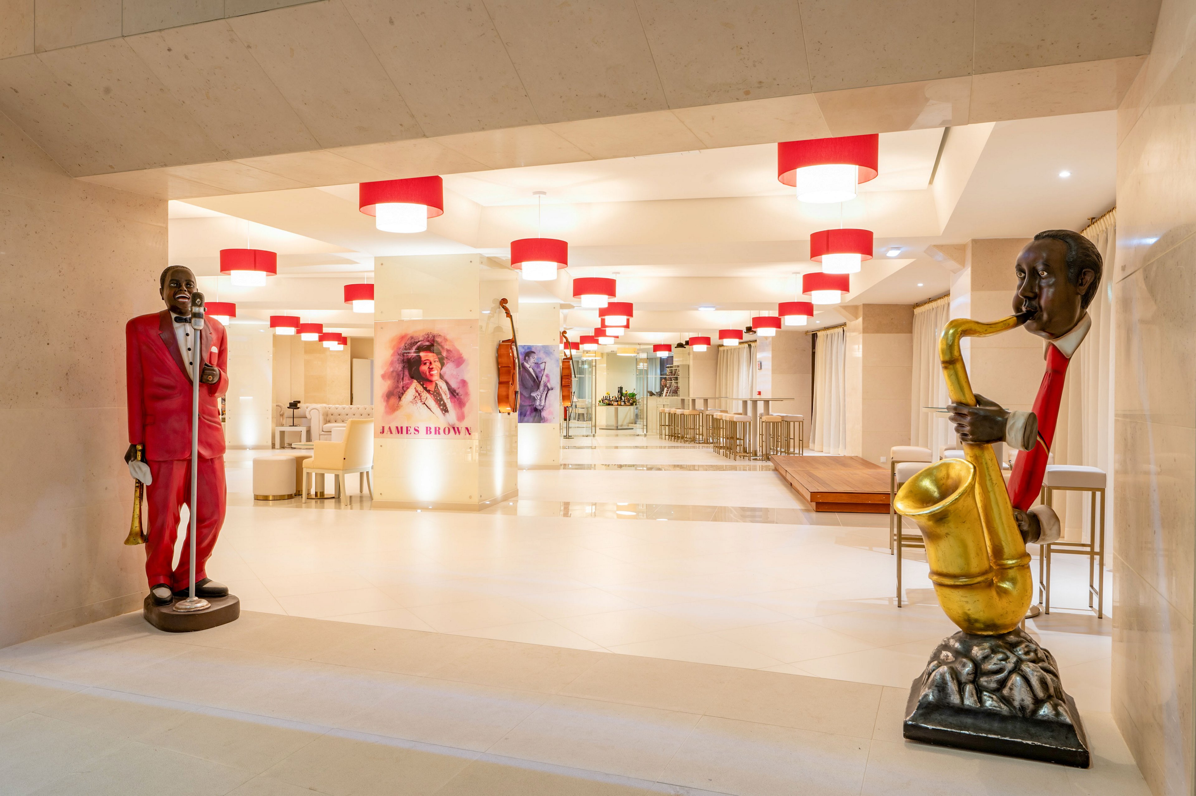 A bright, contemporary lobby features two large sculptures of jazz musicians, one singing into a microphone and the other playing a saxophone. The space is lined with red ceiling lights that create a warm, inviting atmosphere. In the background, colorful jazz-themed artwork decorates the walls, enhancing the musical motif. Clean white floors and neutral walls give the interior a modern, spacious feel.