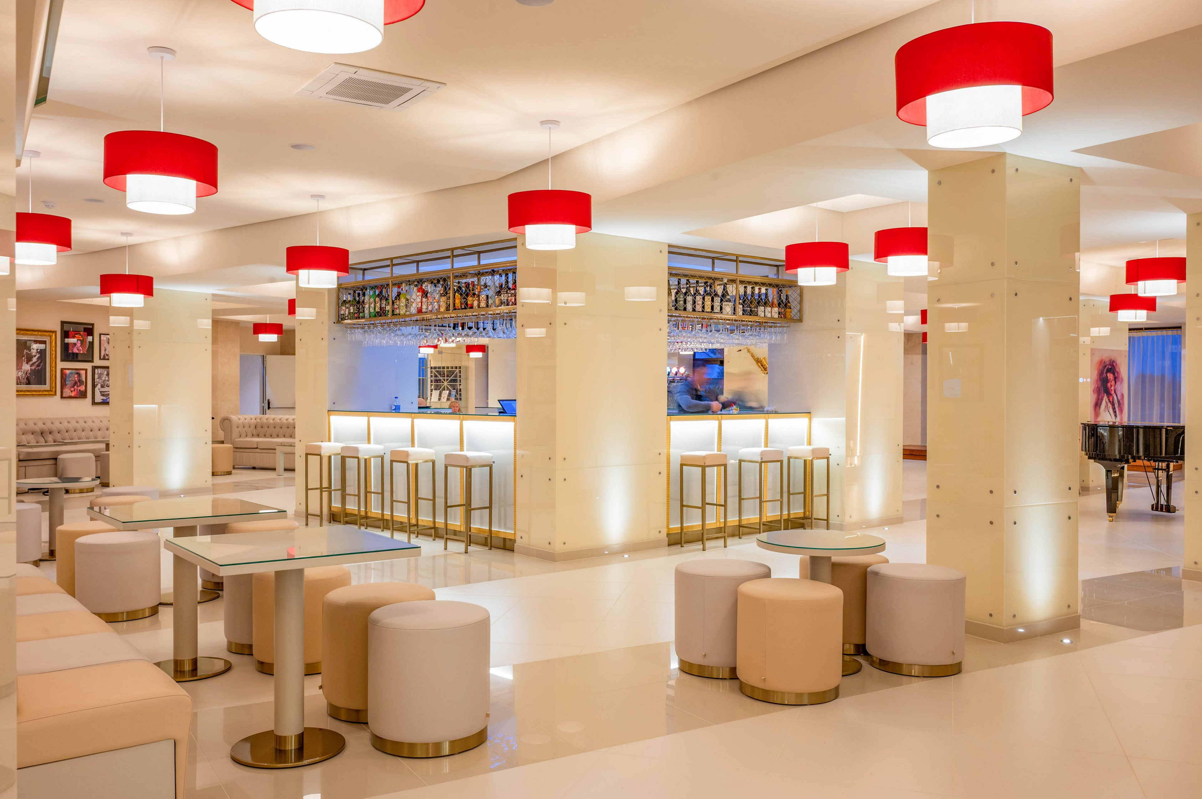 Bright contemporary bar lounge interior featuring glossy white floors and columns with bold red ceiling lights. A central illuminated bar with high stools is surrounded by low ottomans and glass-top tables, creating an open social space. Bottles are displayed along the back bar, and framed artwork decorates the walls, adding visual interest. The overall atmosphere is clean, stylish, and inviting, suitable for hotel guests or upscale events.
