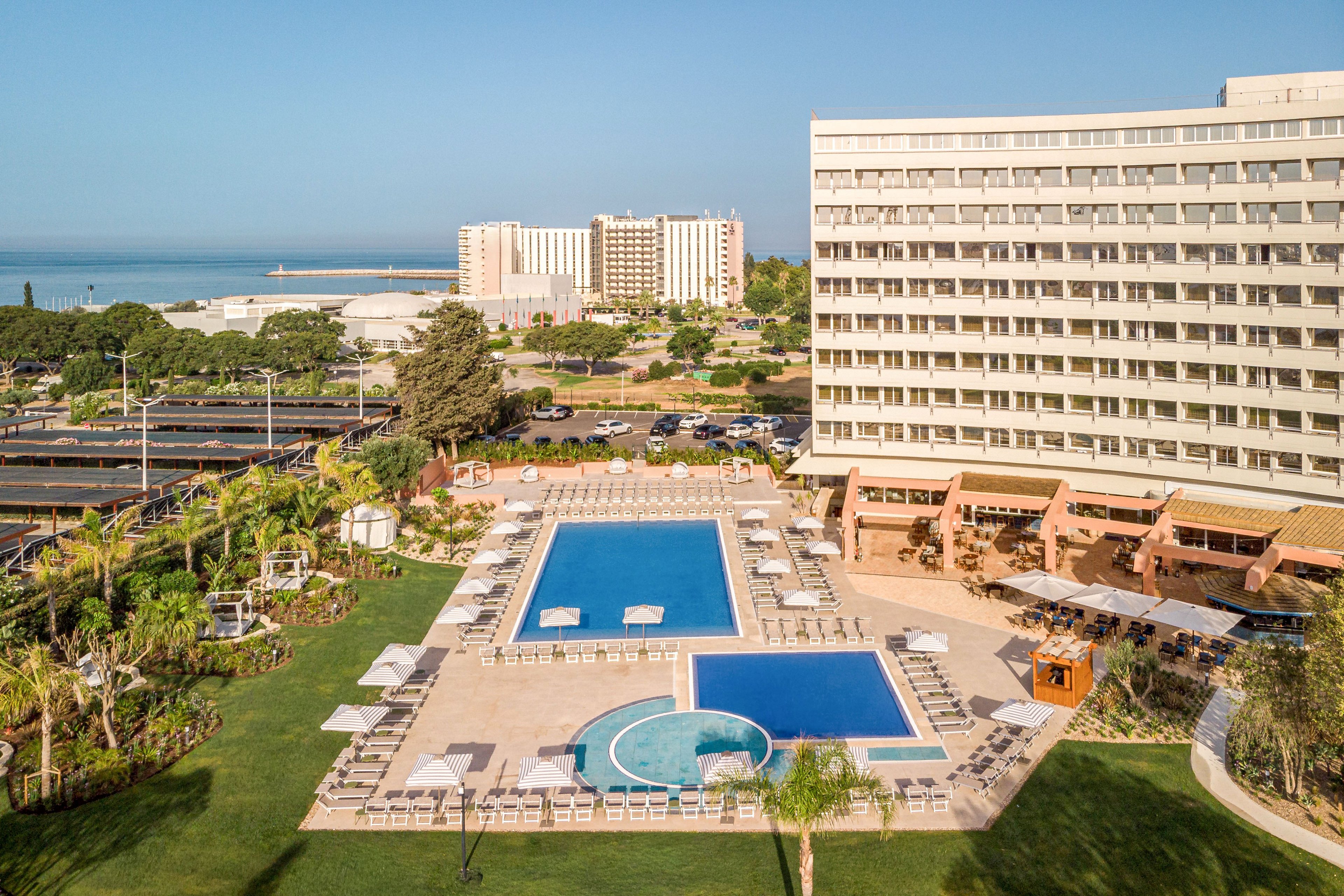 A large multi-story hotel overlooks an expansive outdoor pool area near the sea. The central rectangular swimming pool is surrounded by sun loungers and umbrellas, with a smaller circular pool in the foreground. Lush green lawns, palm trees, and landscaped gardens frame the leisure area, while additional buildings and parking are visible in the background. The scene is captured on a clear, sunny day, emphasizing a relaxed vacation atmosphere.