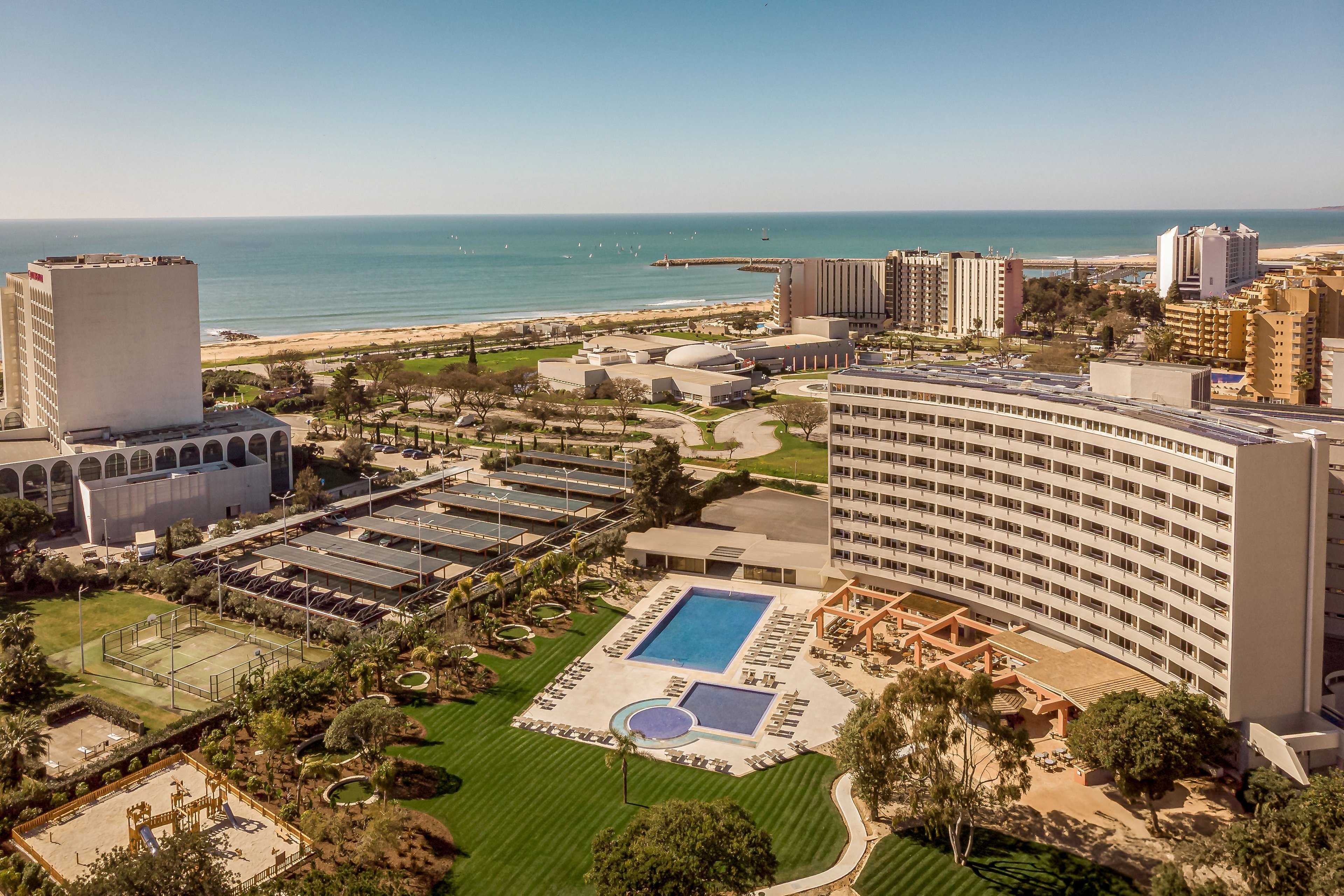 A large seaside resort complex is shown from an elevated viewpoint overlooking the ocean. The scene features a curved multi-story hotel building, a central outdoor swimming pool area, and several tennis courts surrounded by landscaped gardens. Additional high-rise buildings line the beachfront, with a pier extending into the calm sea under a clear sky. The overall atmosphere is sunny and relaxed, highlighting leisure and vacation activities by the coast.