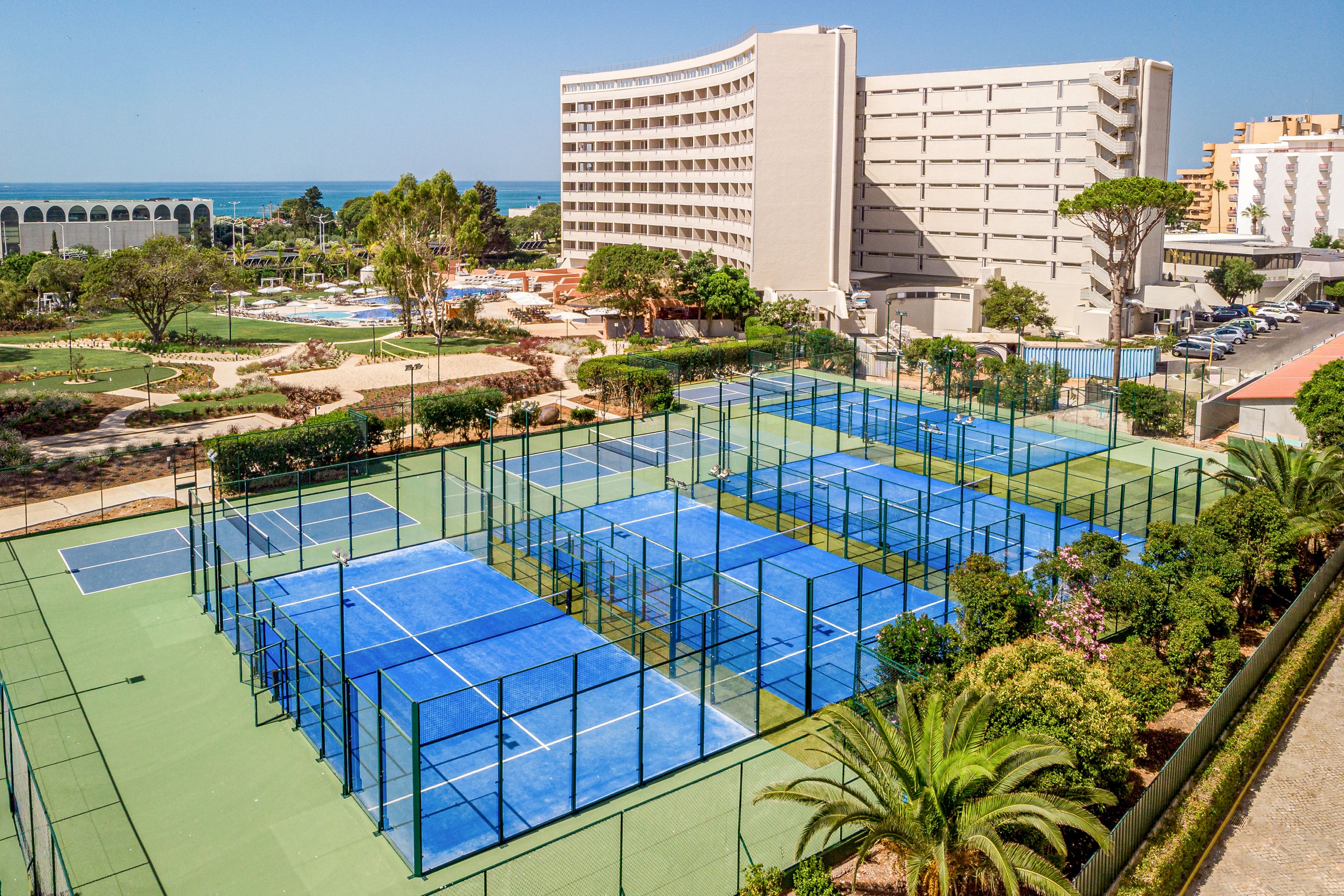 A group of bright blue tennis courts is seen in the foreground of a large seaside resort complex. Tall hotel buildings rise behind the courts, surrounded by landscaped gardens, palm trees, and walking paths. In the distance, additional pools and recreational areas are visible, with the ocean meeting the horizon under a clear sky. The scene conveys a sunny vacation atmosphere focused on outdoor sports and leisure.