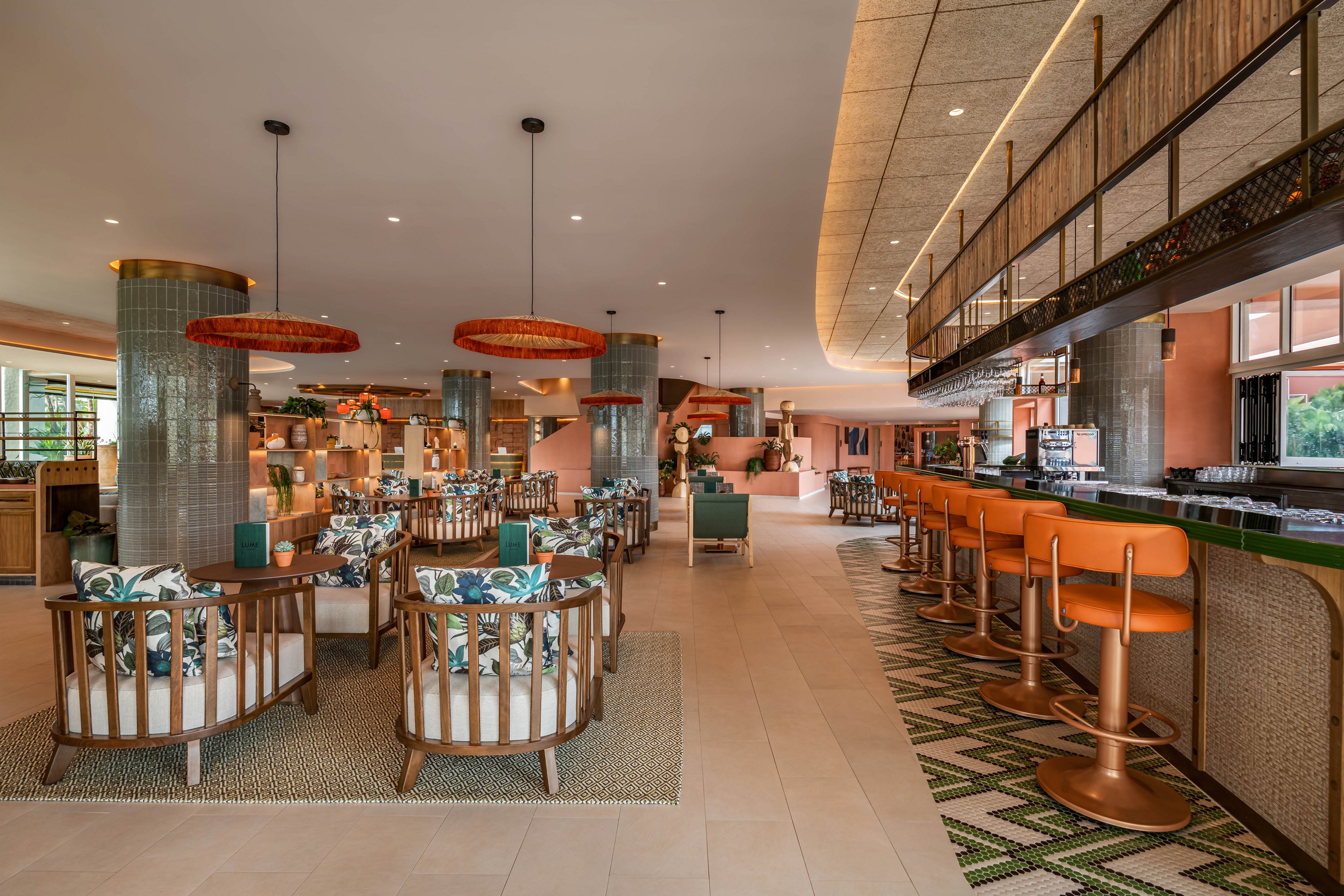 Contemporary hotel bar and restaurant interior featuring warm earthy tones and tropical accents. The space includes a long bar with orange upholstered stools and a patterned green-and-white floor. Lounge-style seating with round wooden chairs and botanical print cushions fills the open dining area. Large pendant lights, textured columns, and expansive windows create an inviting, resort-like atmosphere.