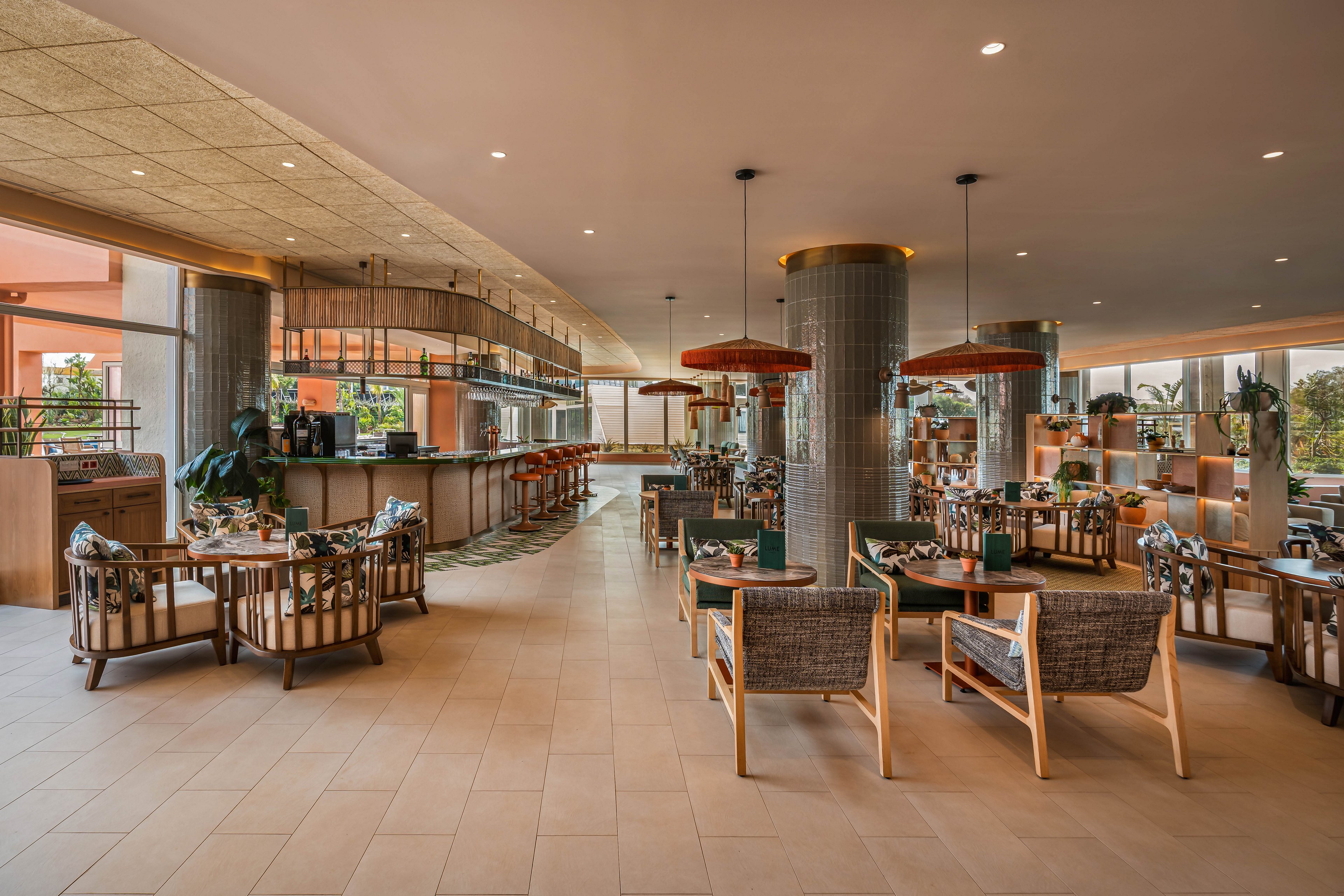 Spacious contemporary restaurant interior featuring a central bar and multiple seating areas. Wicker and upholstered chairs surround wooden tables arranged across a tiled floor. Large columns, hanging light fixtures, and warm neutral tones create a relaxed, upscale atmosphere. Floor-to-ceiling openings reveal greenery and natural light from the outdoor area.